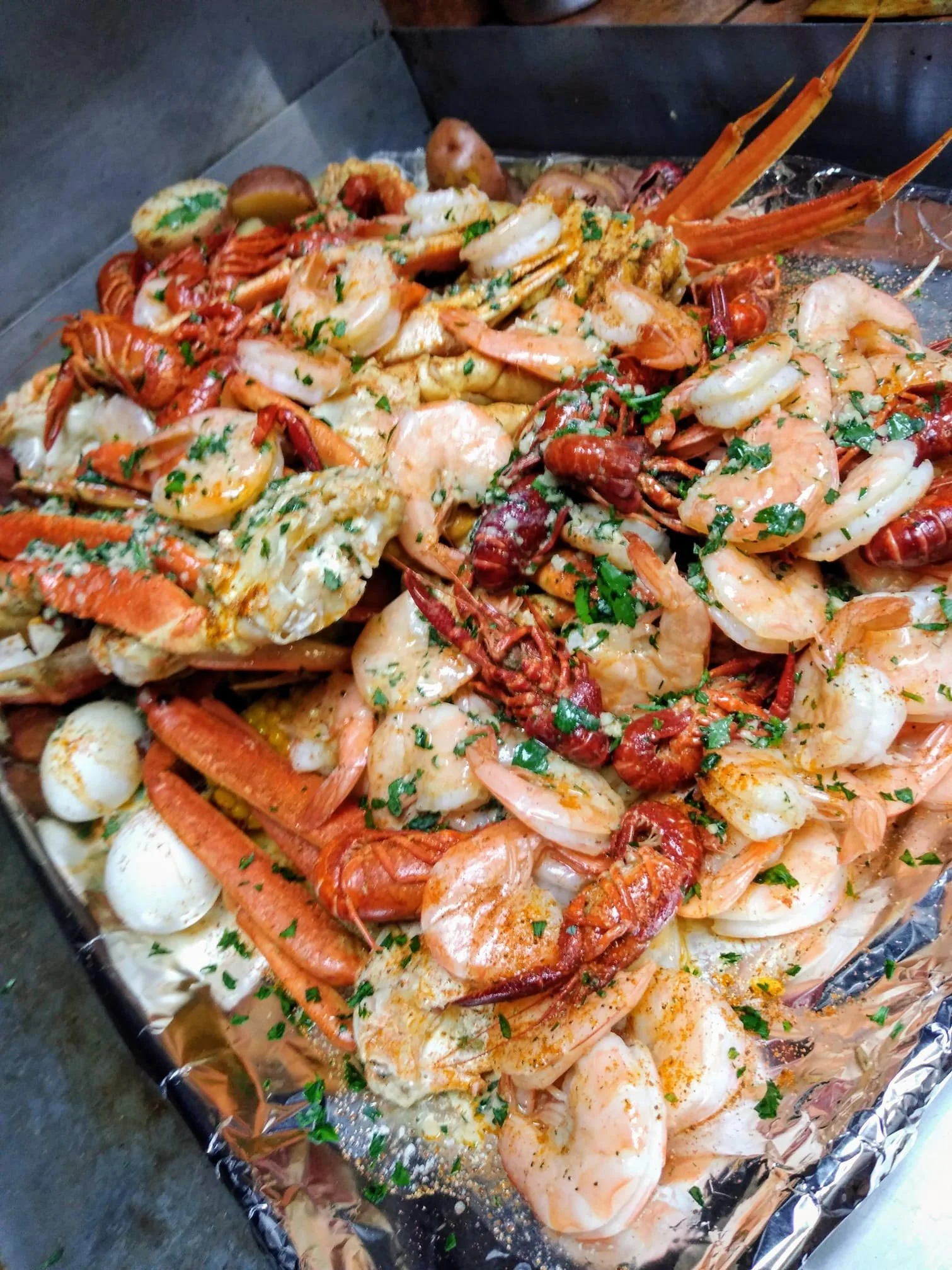 A tray of cooked seafood including shrimp, lobster, crawfish, and boiled potatoes, garnished with chopped parsley.