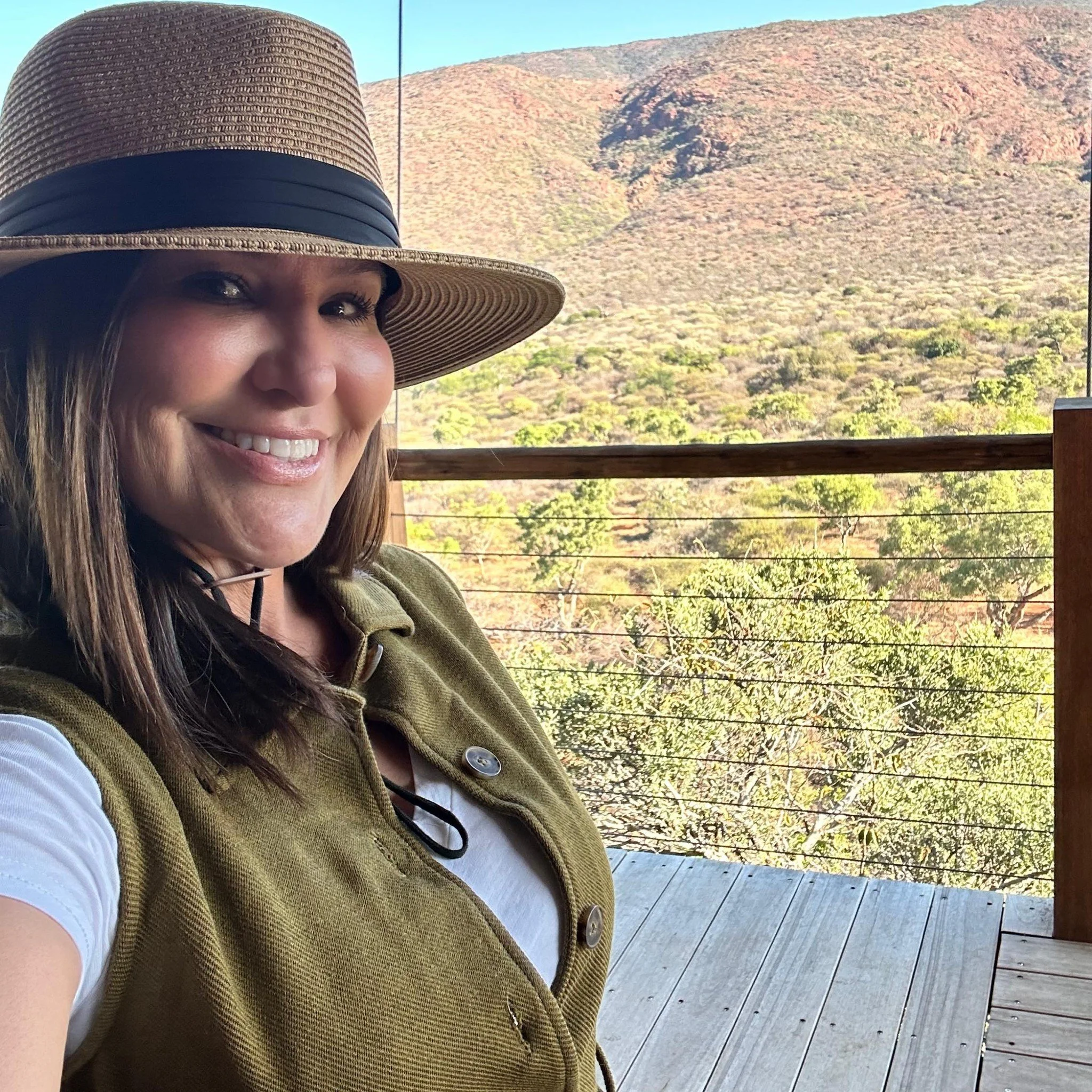 A smiling woman wearing a wide-brimmed hat and an olive-green vest takes a selfie on a wooden balcony with a scenic view of a hillside covered in sparse vegetation and trees.