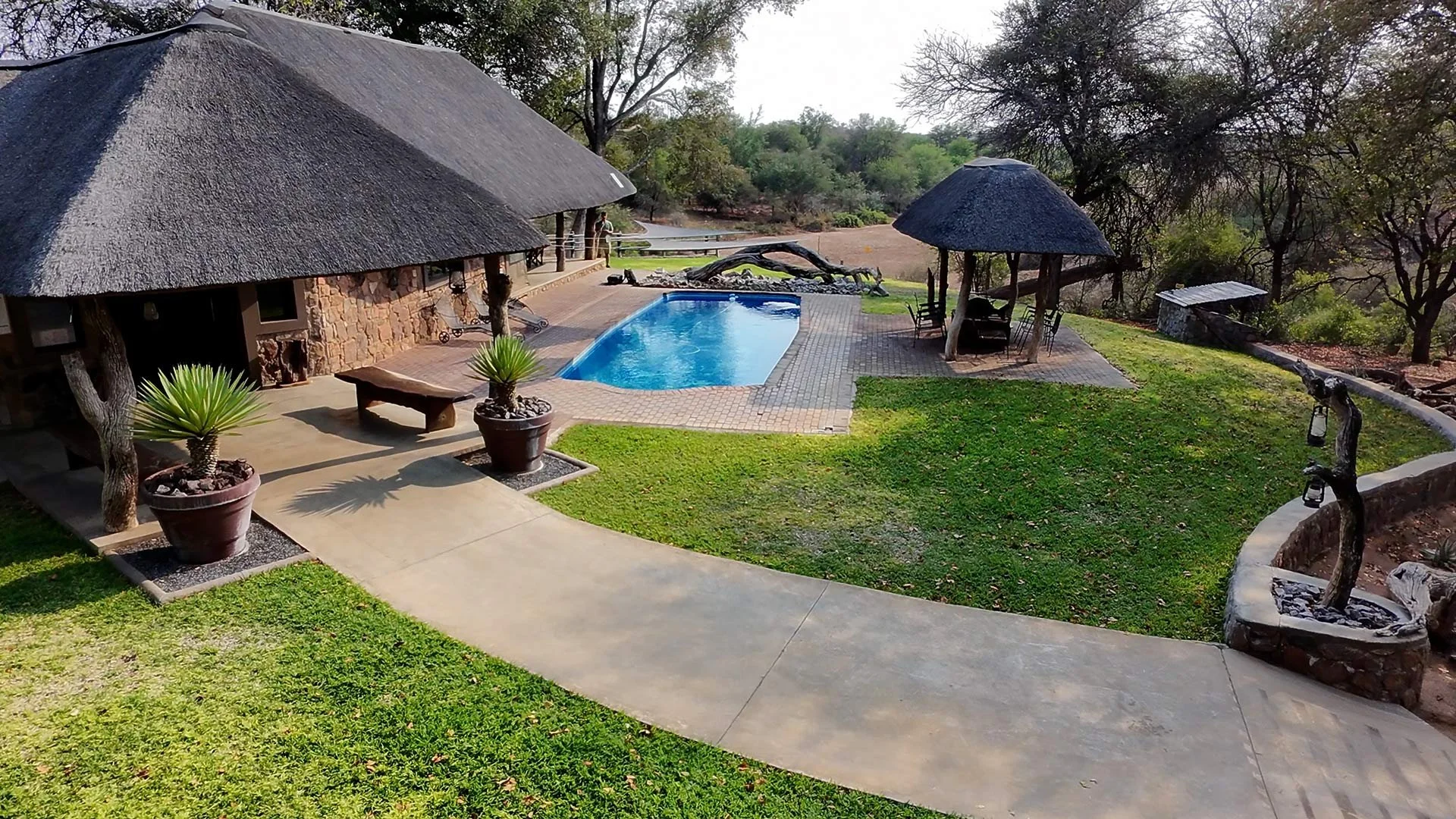 A backyard with a swimming pool, two thatched-roof gazebos, and outdoor seating surrounded by trees and greenery.