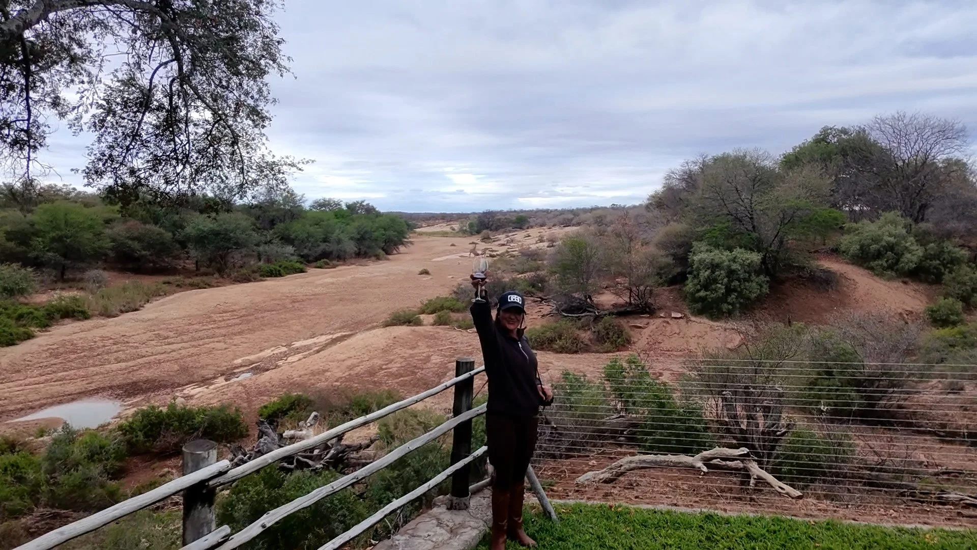 A woman standing on a wooden platform with a safety railing, holding up a glass, in a natural landscape with sparse trees, bushes, and rock formations, overcast sky, and distant animals on the horizon.