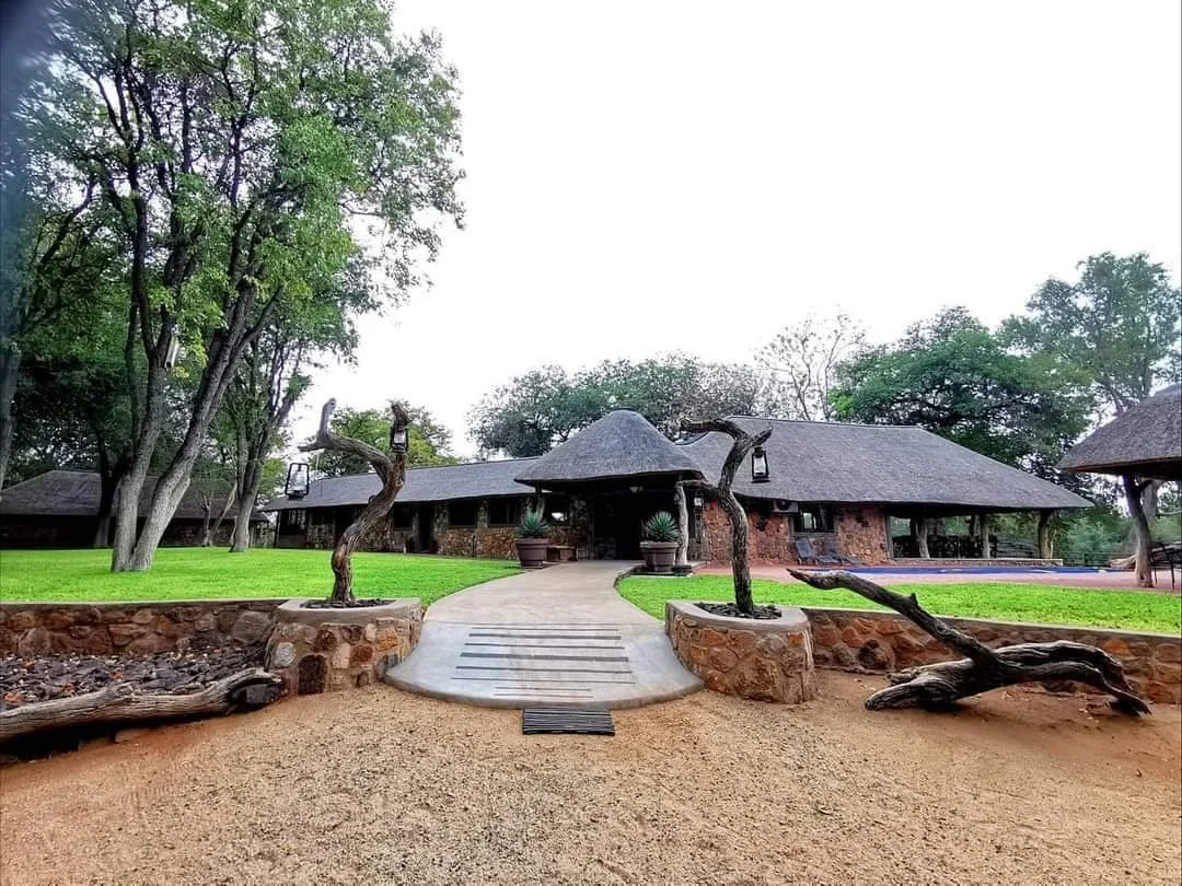 A large building with a grey, thatched roof, brick walls, and a wide porch, surrounded by green grassy yard and large trees. There is a curved pathway leading to the entrance and decorative twisted tree trunks on stone bases in the foreground.
