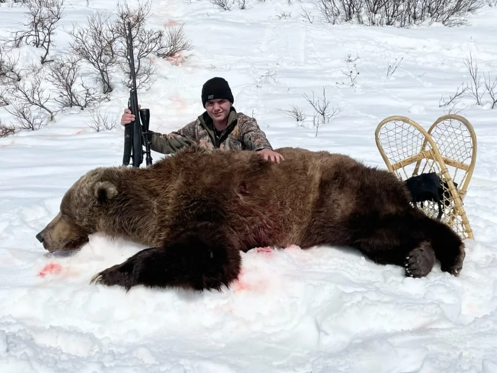 A young boy in camouflage clothing and a black beanie kneels next to a large dead bear lying on its side in the snow. The boy holds a rifle, and there are two snowshoes behind the bear in the snow. Bare trees are visible in the background.
