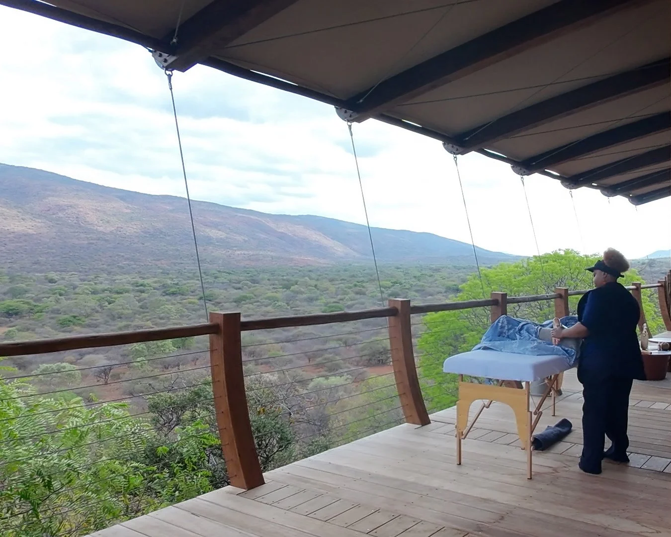 Outdoor massage session on a wooden deck overlooking a vast green valley and mountain range, with a therapist standing beside a client under a shaded canopy.