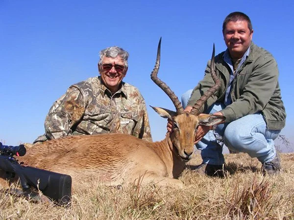 Two men crouching next to a large antelope with twisted horns, lying on dry grass in a field under a clear blue sky.