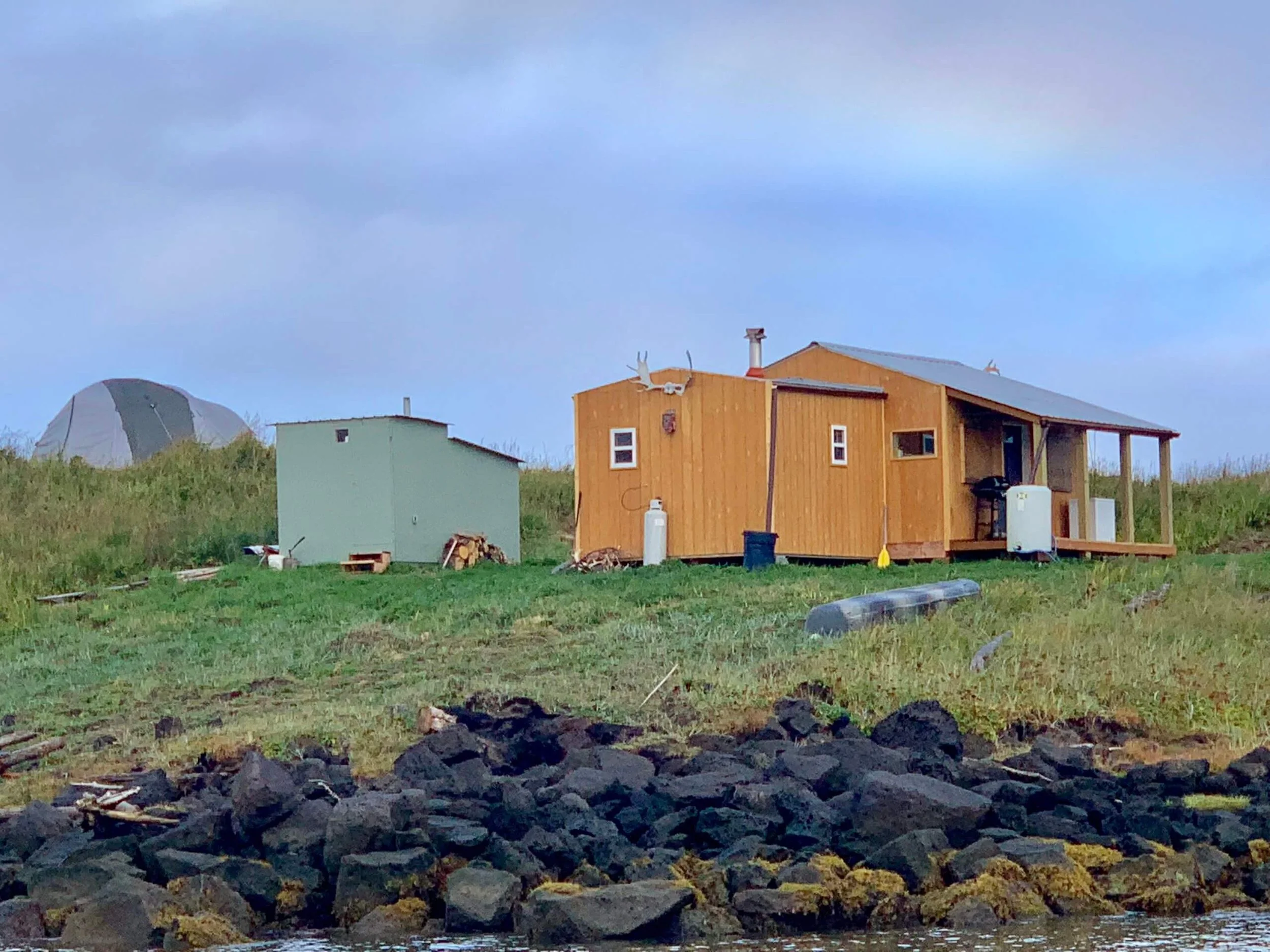 Coastal scene with a small wooden house on grassy land, a separate small green shed, and a dome-shaped tent in the background, along rocky shoreline and cloudy sky.