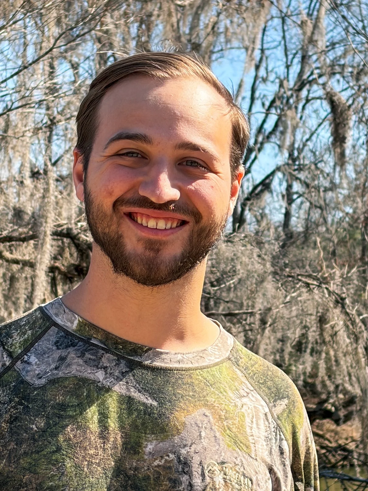 A young man with fair skin, dark neatly combed hair, wearing a black tuxedo and white dress shirt, smiling in a portrait photo with a spanish moss covered tree background.