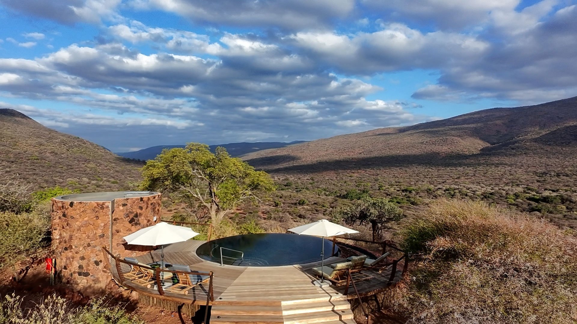 A round infinity pool on a wooden deck with lounge chairs and white umbrellas, overlooking a valley with mountains and sparse vegetation under a partly cloudy sky.
