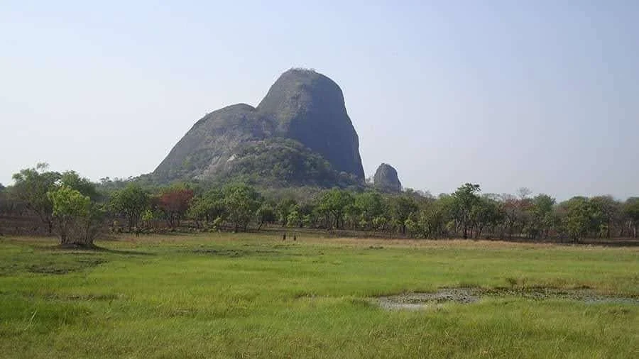 A white pickup truck driving along a rugged dirt path through a vast, dry African landscape with sparse bushes and trees stretching toward the horizon.