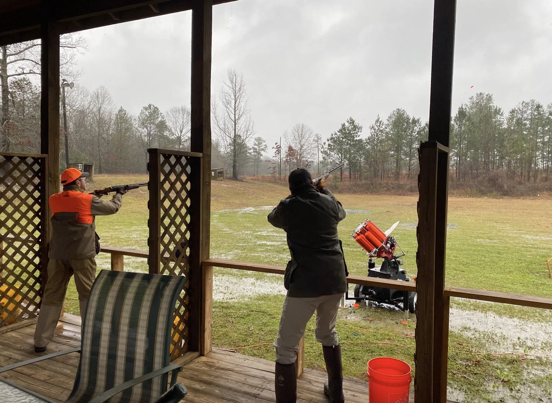 Two people shooting at a range from a covered wooden platform, with one person aiming a rifle and the other aiming a shotgun, with cloudy weather and trees in the background.