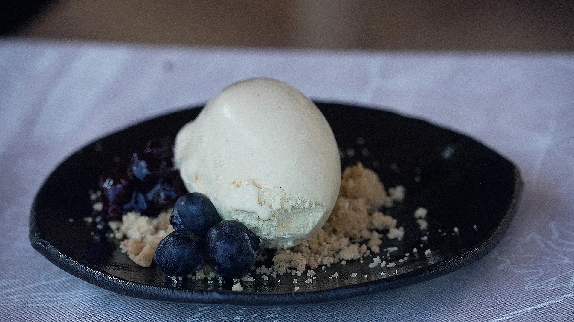 A scoop of vanilla ice cream with blueberries, blueberry compote, and crumbled cookie on a black plate.