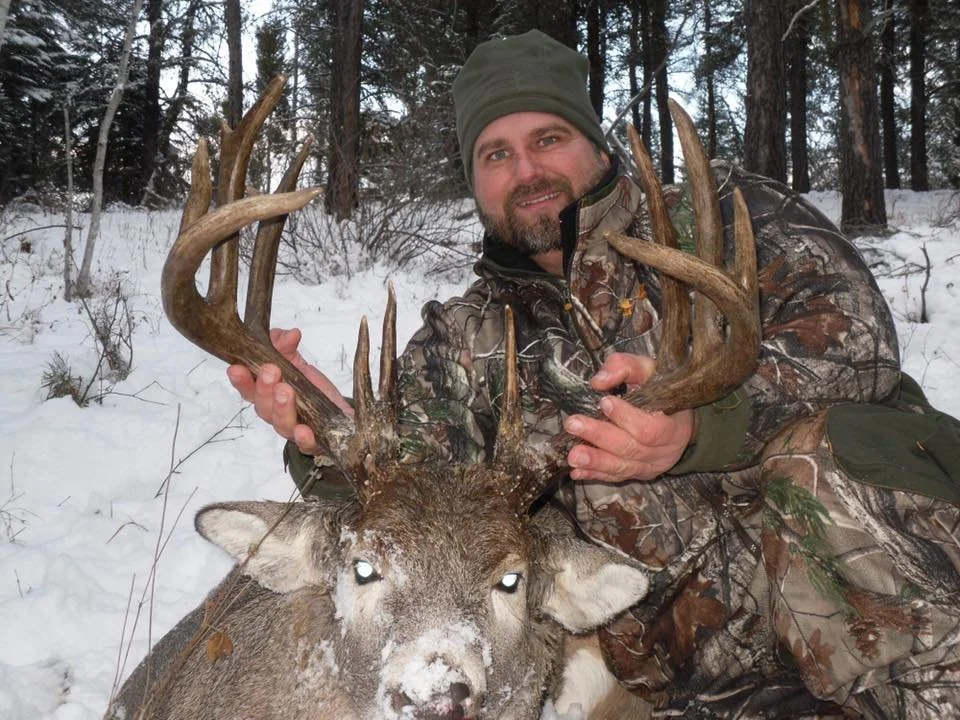 John Wilson in camouflage and green winter gear holding the antlers of a large deer with antlers in a snowy forest.