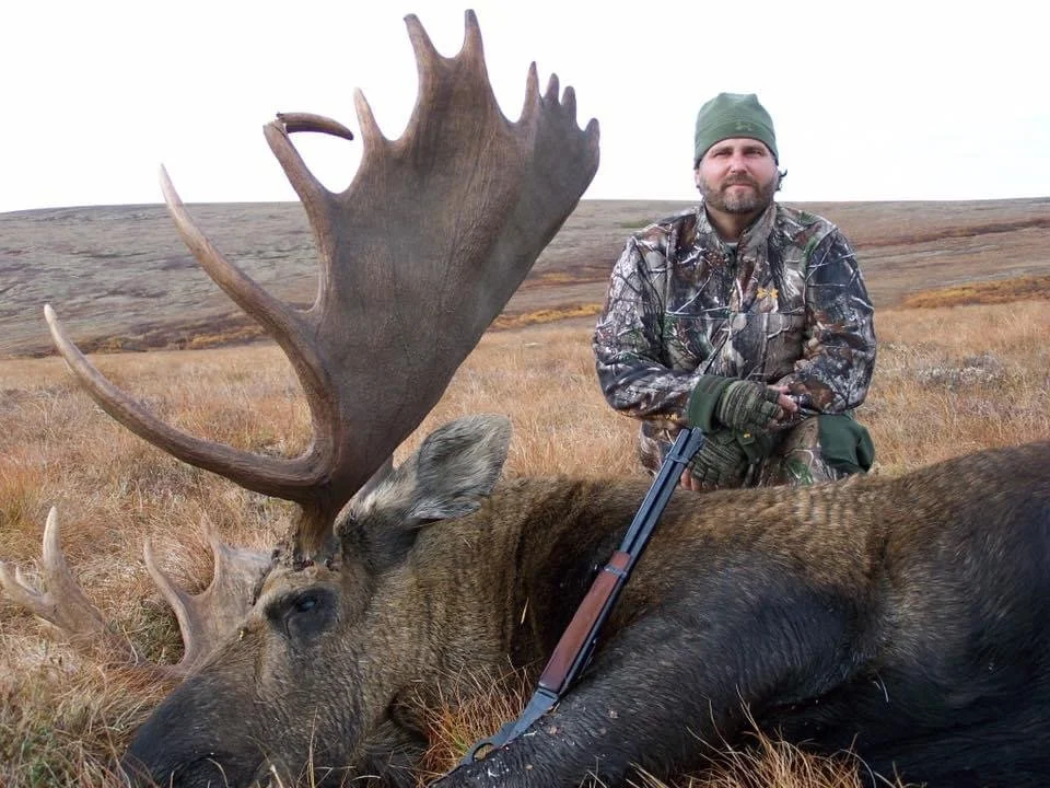 A man in camouflage clothing kneels next to a large dead elk with big antlers, holding a rifle, in a grassy field with rolling hills in the background.
