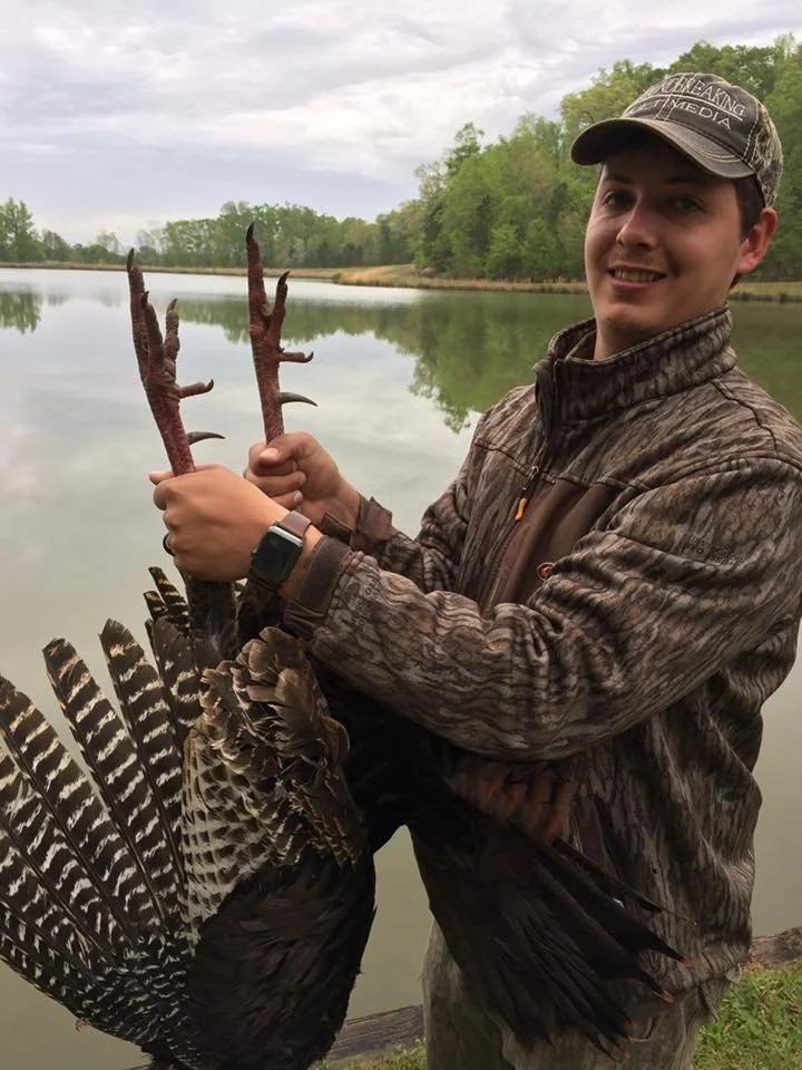 Young man standing by a lake holding a pair of turkey legs with a wild turkey's tail feathers in his other hand, wearing camouflage clothing and a cap, with trees and water in the background.