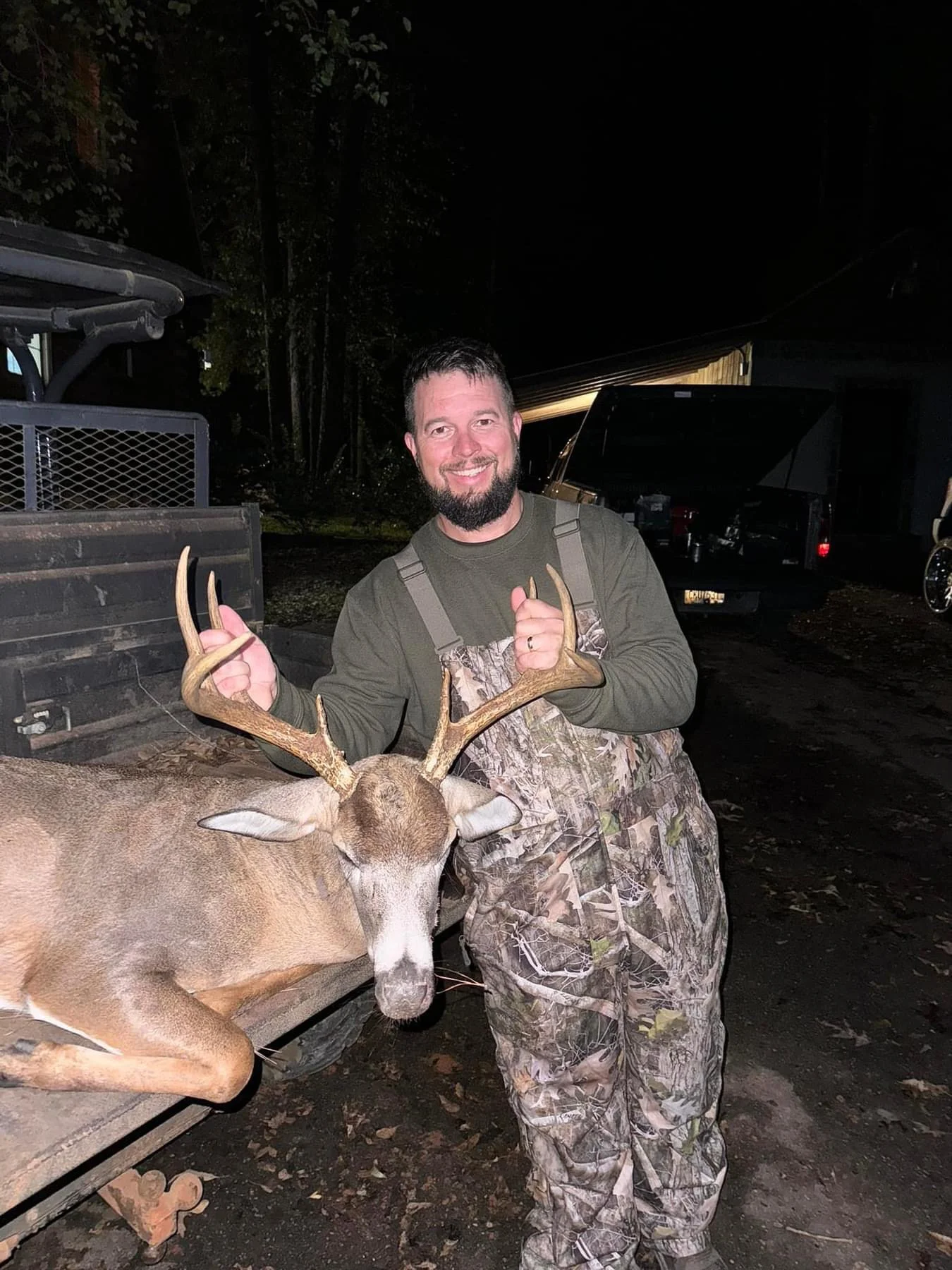 A man in camouflage clothing smiling and holding the antlers of a deer he has presumably hunted, standing outdoors at night.