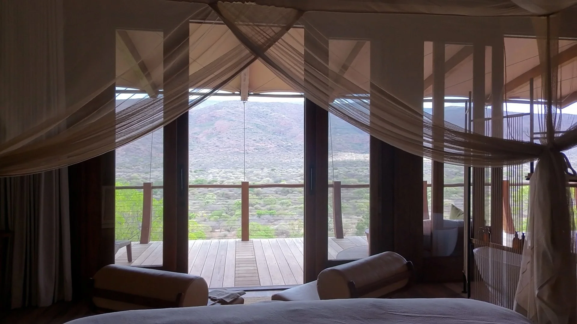 View from a luxury safari bedroom looking out through large glass doors onto a wooden deck overlooking a vast valley and distant mountains, framed by soft canopy netting.