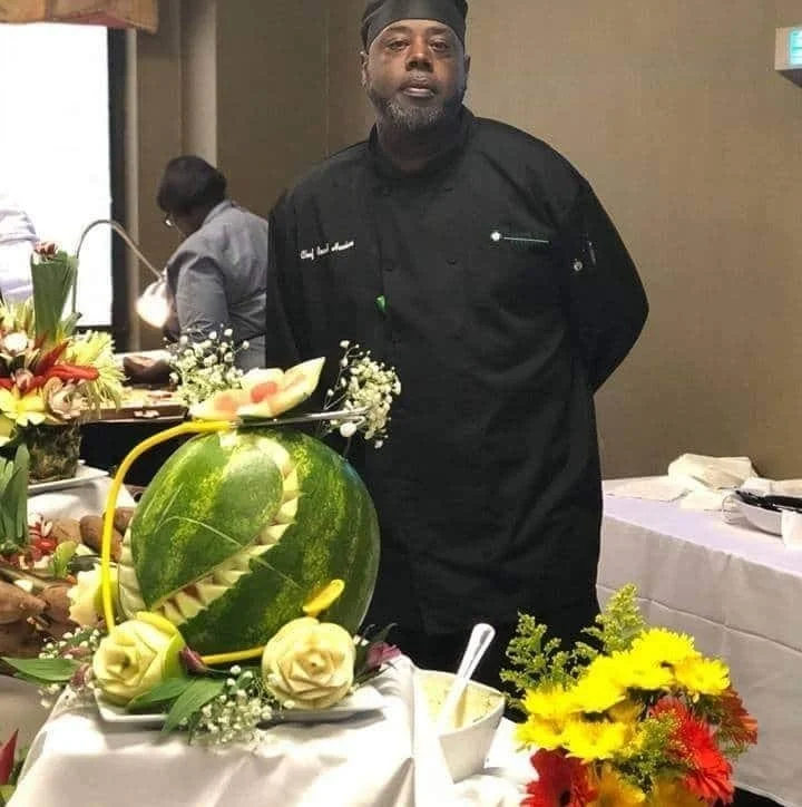 A chef in a black uniform standing behind a table with a carved watermelon centerpiece and various floral arrangements.