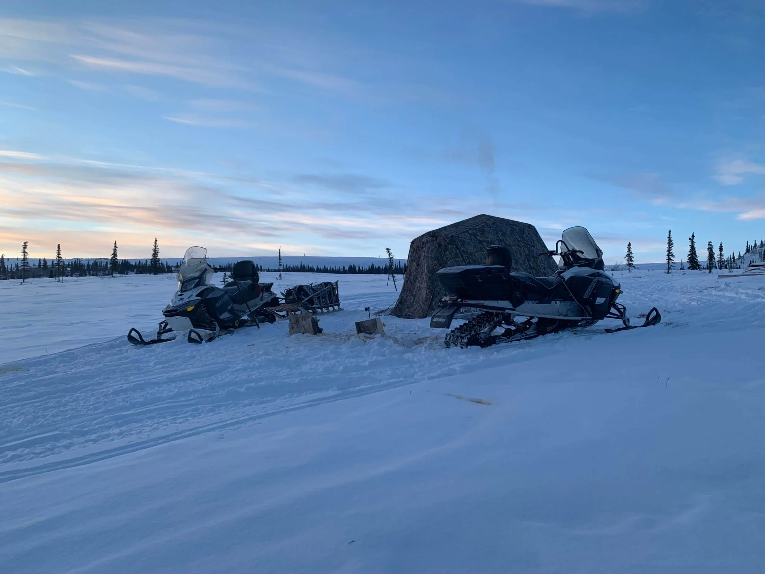 Two snowmobiles parked in a snowy landscape under a blue sky with clouds, with a tent and trees in the background.