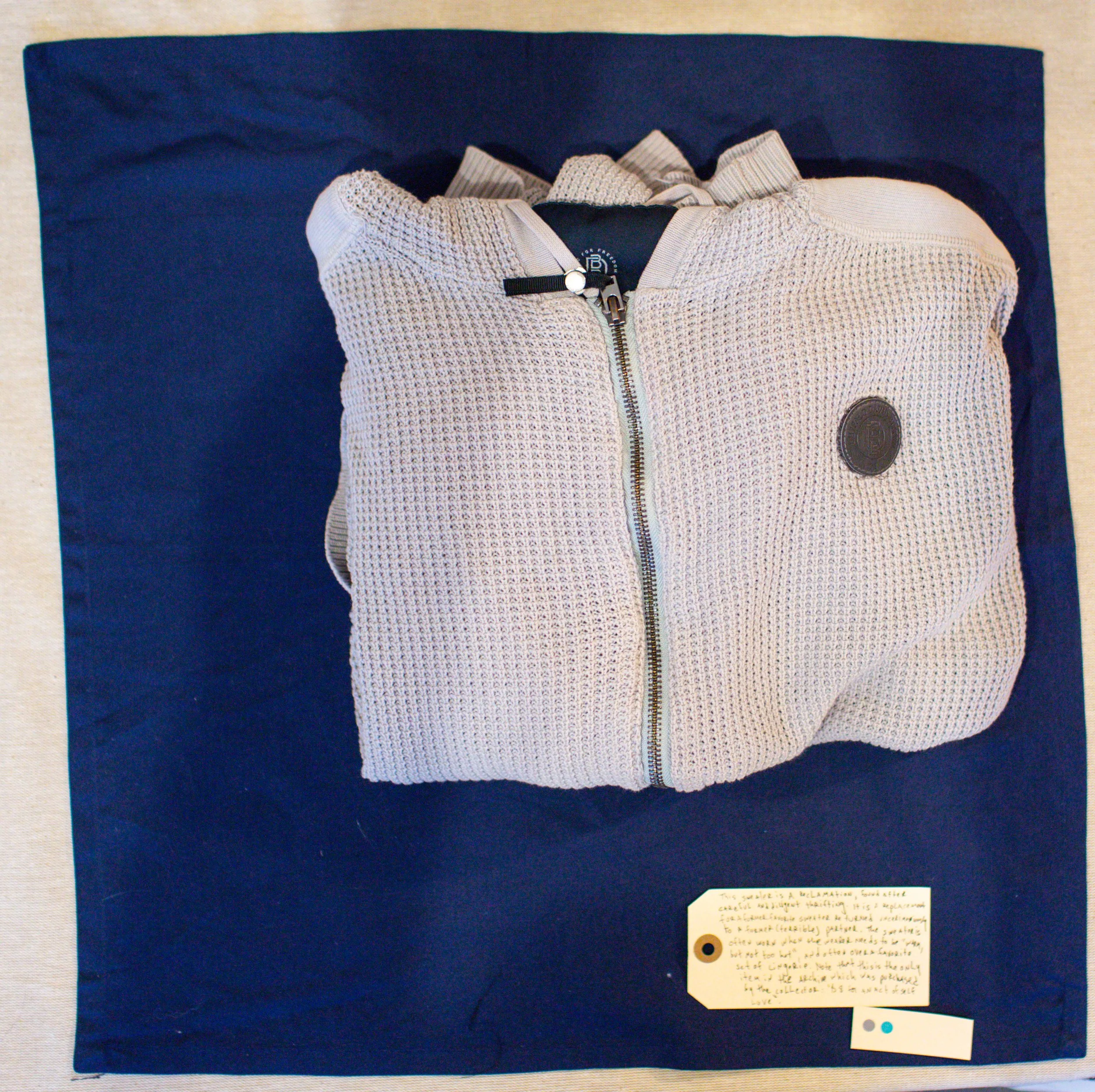 A beige zip-up sweater with a circular logo on the left chest, displayed on a dark blue background, with a small handwritten note attached at the bottom.