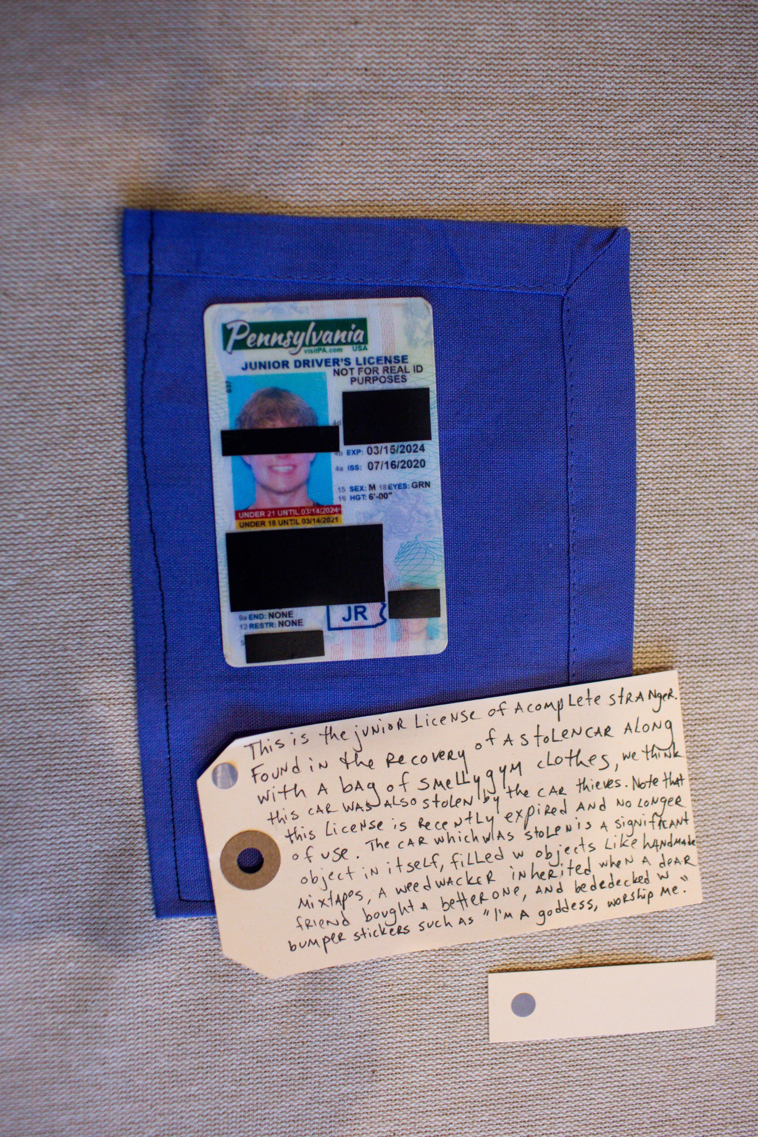 A Pennsylvania junior driver's license in a blue sleeve, accompanied by a handwritten note explaining it was found with a bag of smelly gym clothes and car thefts. The note describes the license as expired, no longer in use, and details the discovery