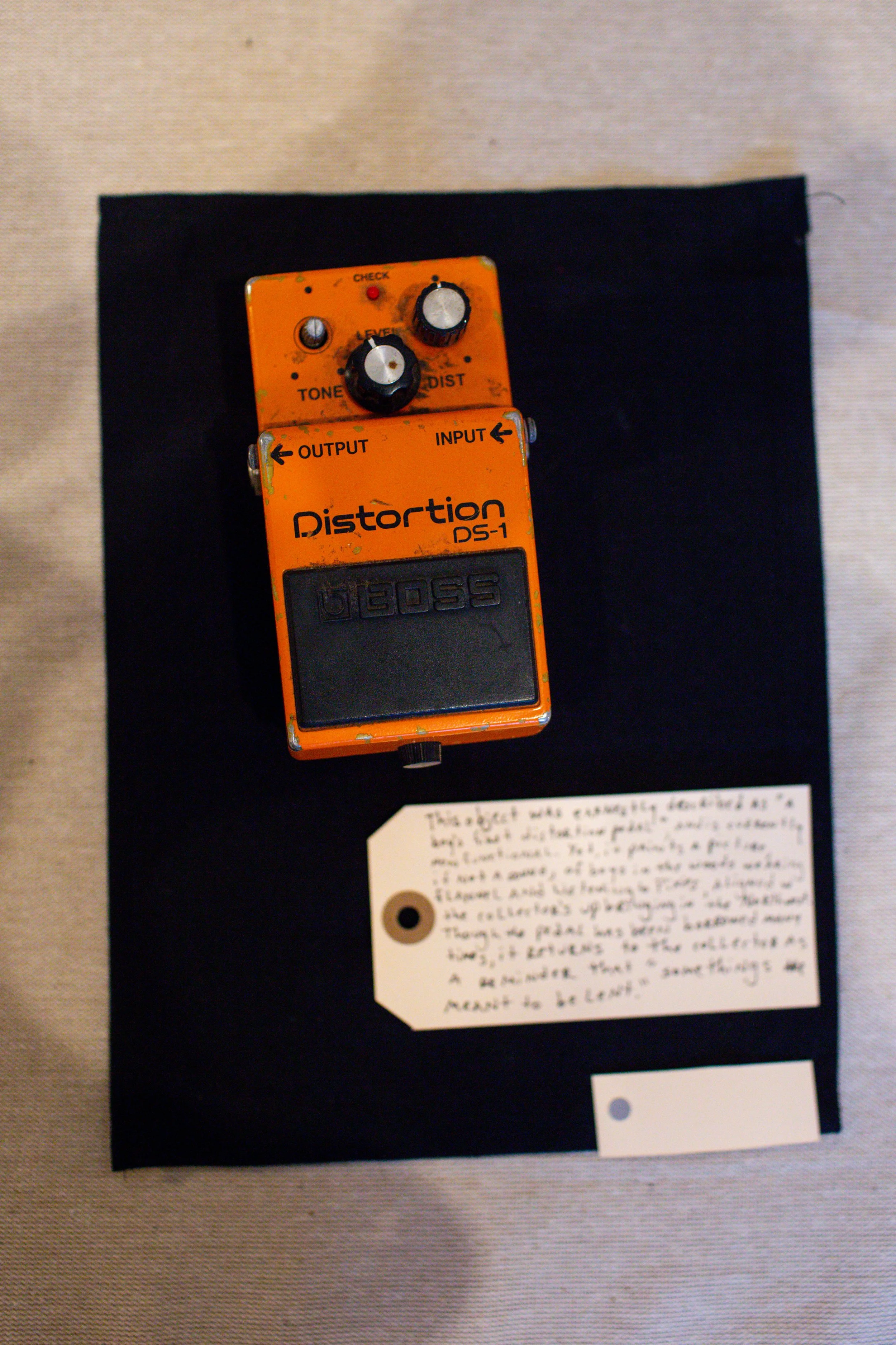 A Boss DS-1 distortion guitar pedal, orange with black rubber foot switch, placed on a black cloth, with a handwritten note below it.