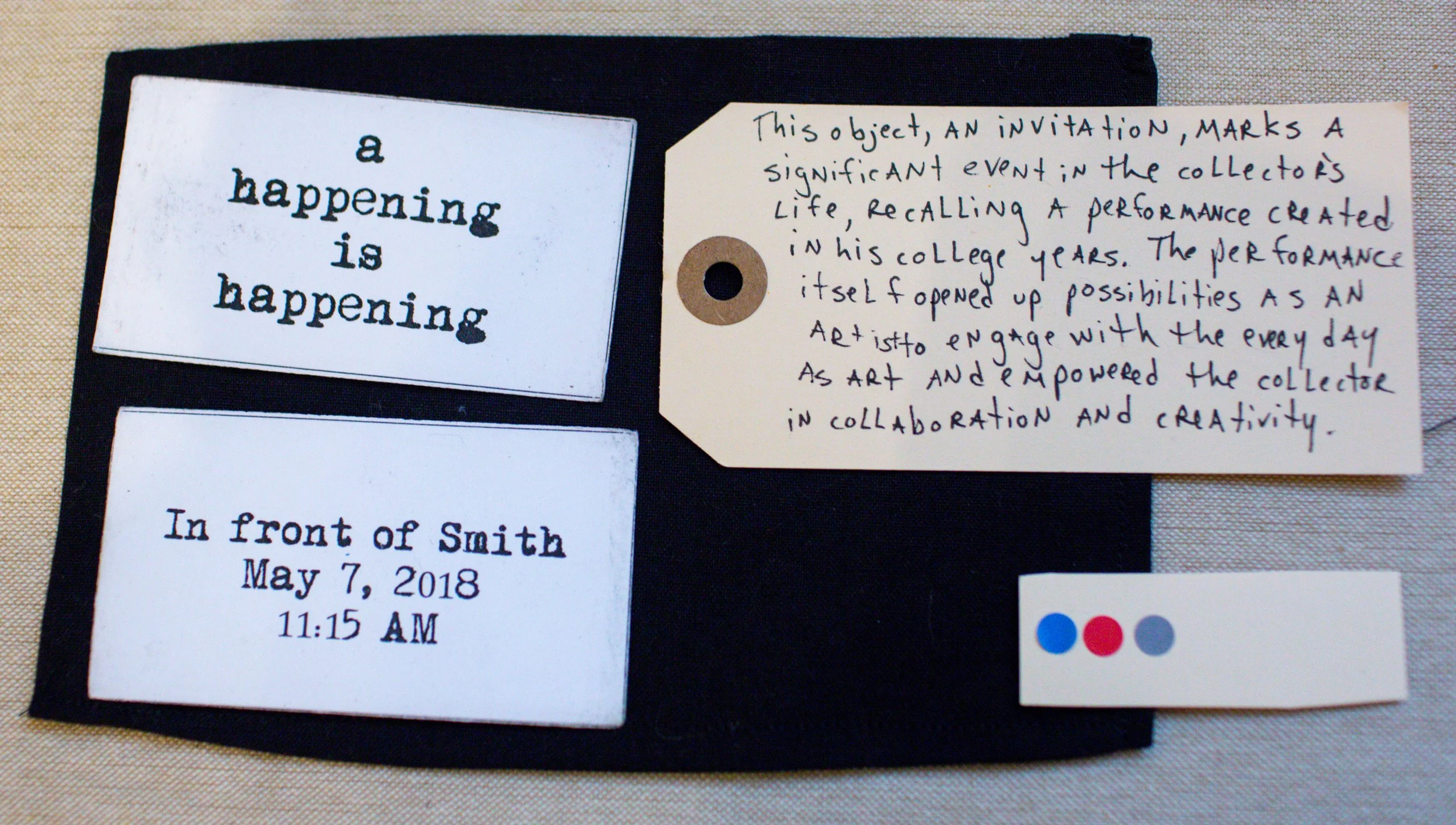 A black fabric background with three items: a white label with black text reading "a happening is happening," a white event details card, and a beige handwritten note on a paper tag discussing the significance of the event. There is also a small whit