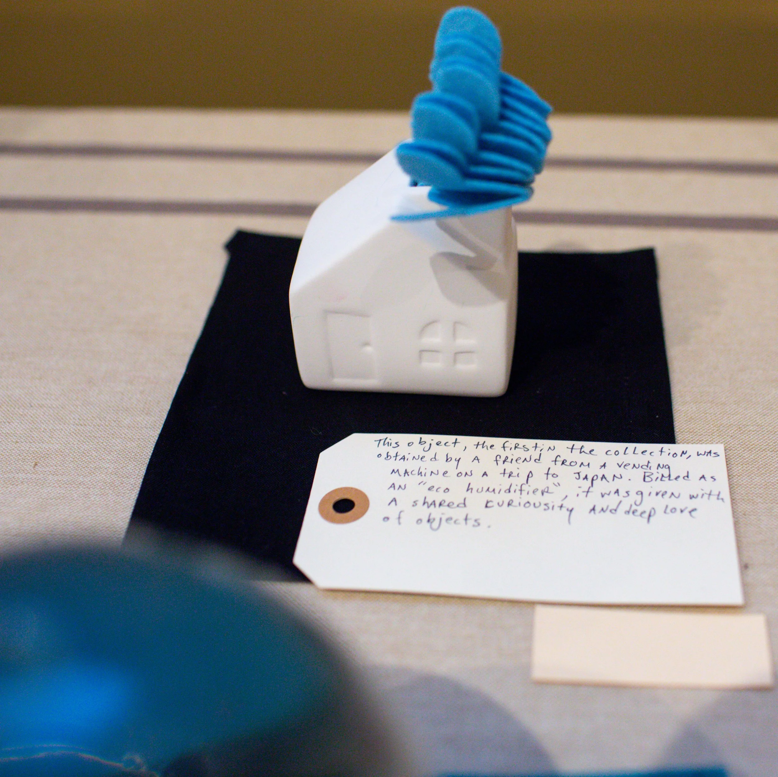 A small white ceramic house-shaped object with a blue spiral object on top, placed on a black cloth on a table. A handwritten note describes the object as an 'eco humidifier' made from a collection obtained from a friend.