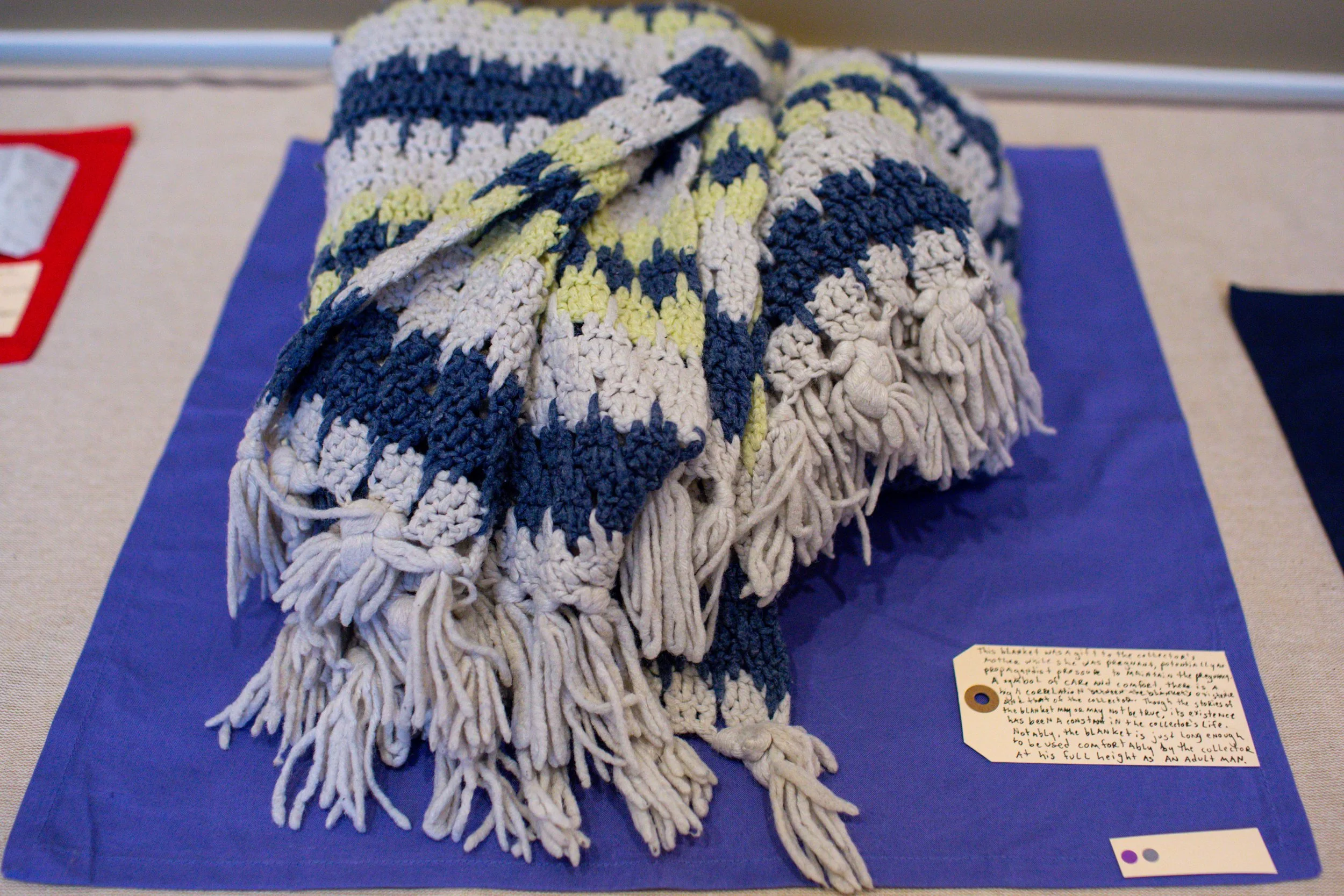 A large, handmade knitted blanket with a pattern of blue, white, and yellow, draped over a blue cloth on a display table.