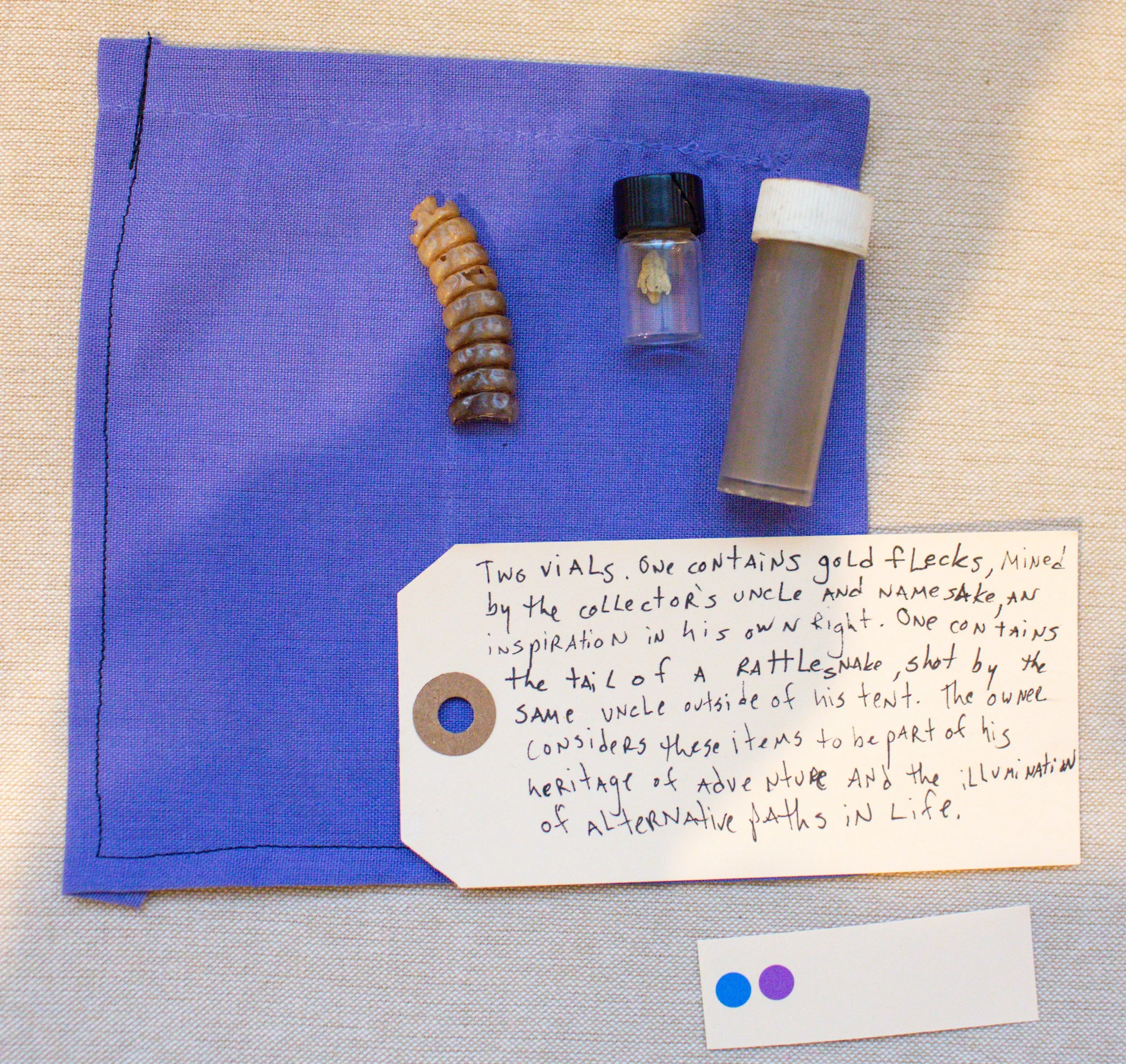 A blue cloth with a small jar containing a tiny object, a taller cylindrical container, and a caterpillar-like object, alongside a handwritten note explaining that the items are part of a collection of vials with personal significance, including a ra