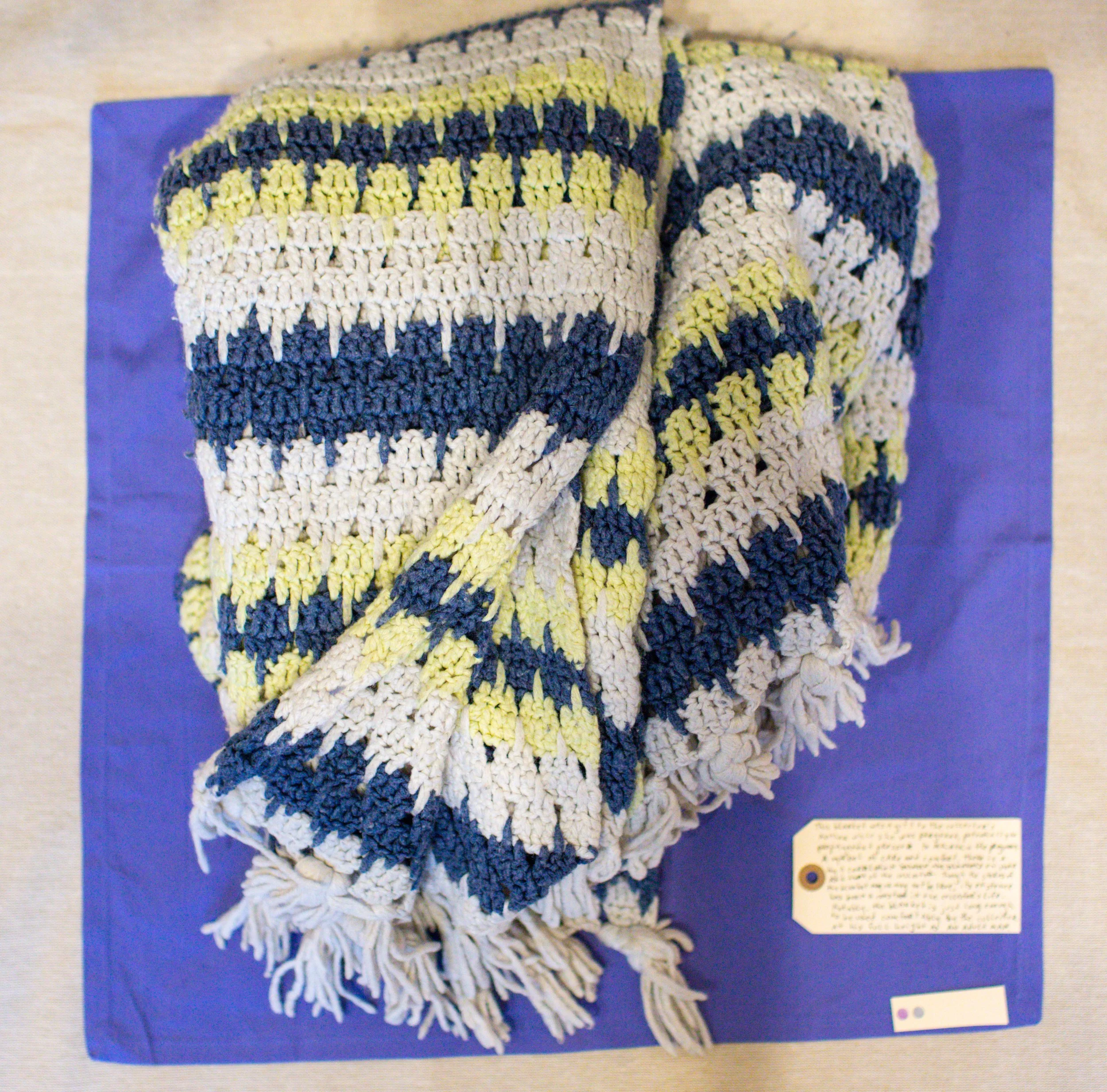 A folded, striped, multicolored knitted blanket with fringe on a blue background and a small informational plaque at the bottom right.