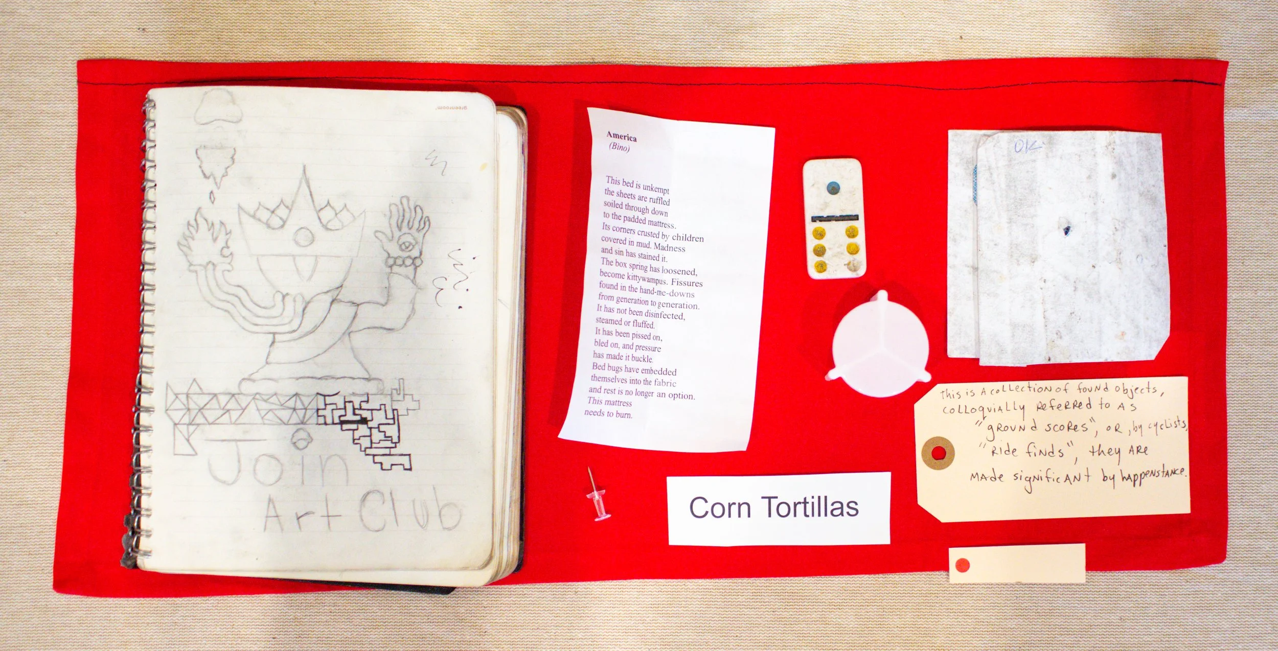 A red display board containing a notebook with sketches, a printed poem, and handwritten notes about objects collected for an art project, labeled 'Corn Tortillas.'