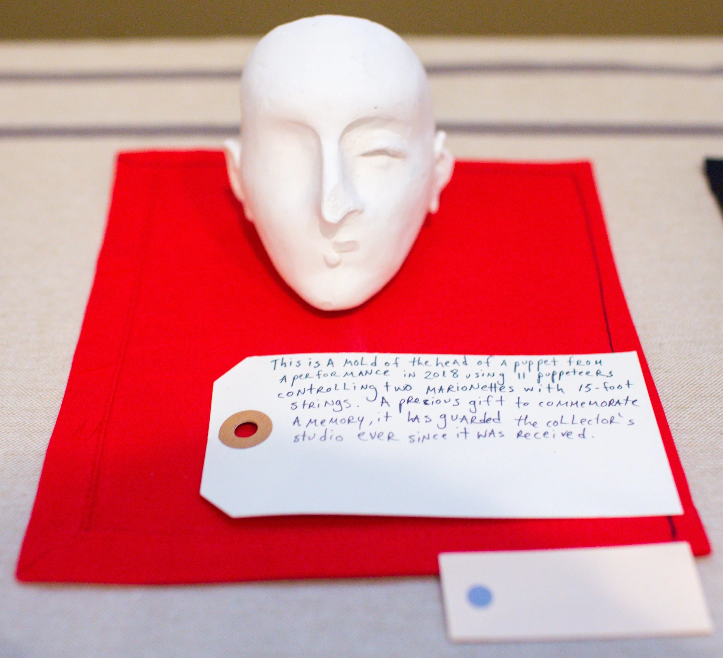 White molded sculpture of a puppet head placed on red fabric with a handwritten note explaining its significance.