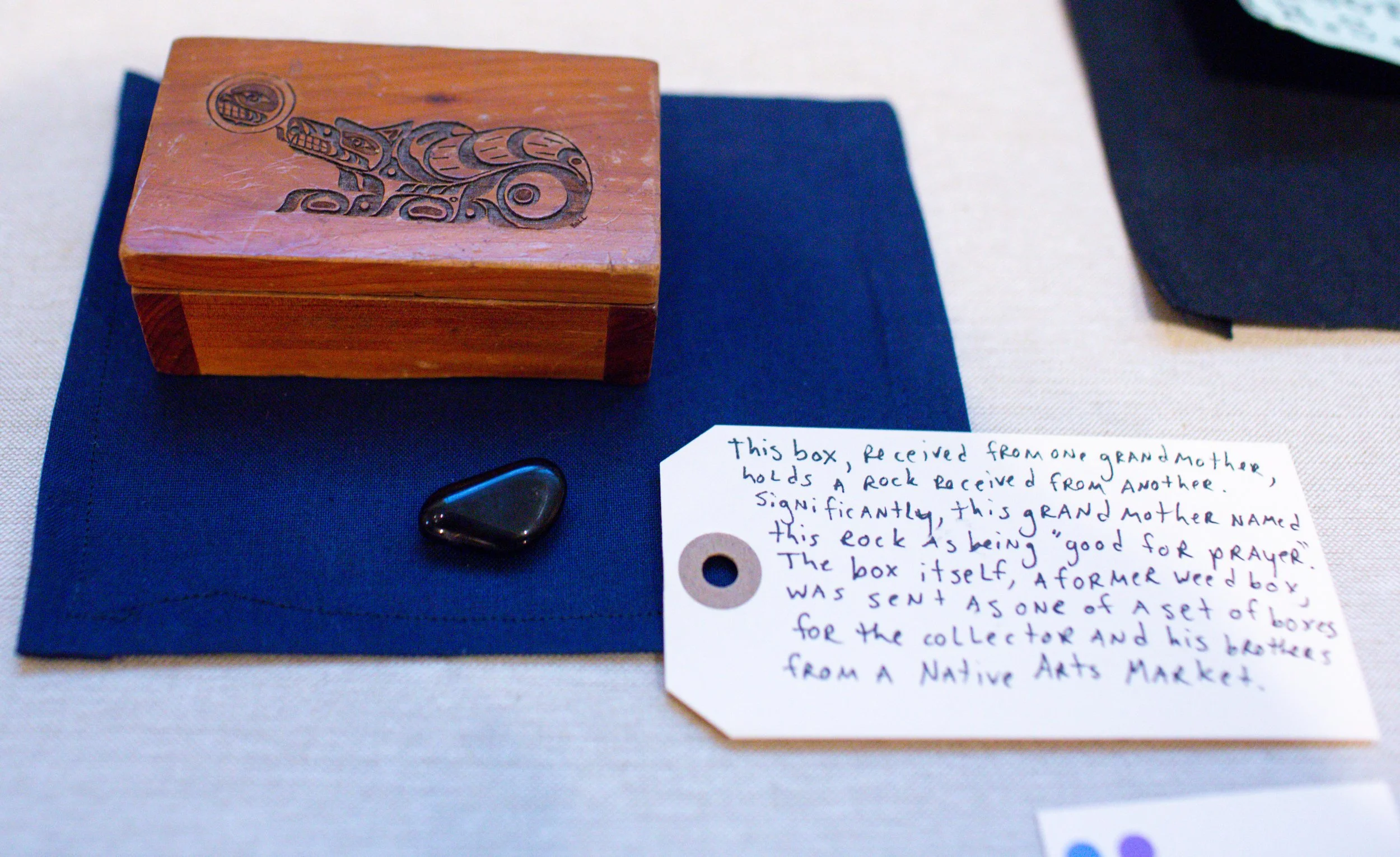 A wooden box with Indigenous art carved into the top, a black smooth stone, a blue felt cloth, a handwritten note describing the items, and part of a dark fabric are displayed on a light-colored surface.