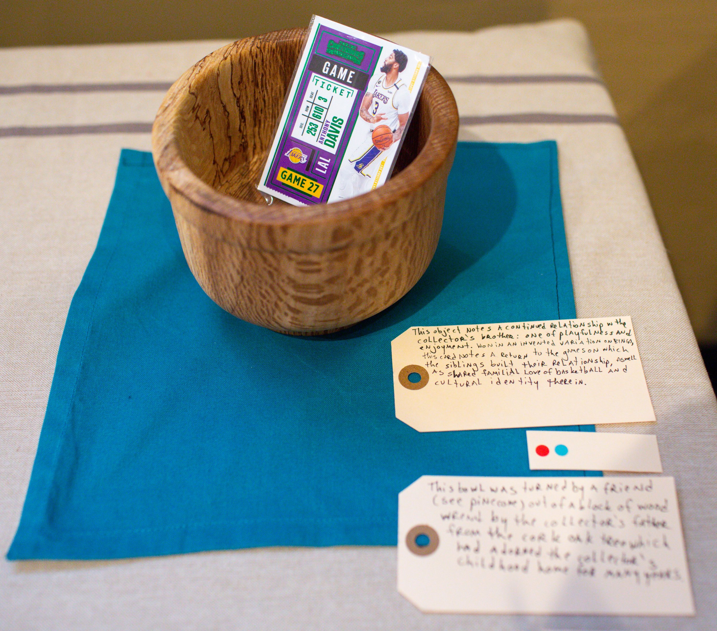 A wooden cup with a Lakers game ticket inside, placed on a turquoise cloth on a beige surface. There are two handwritten notes beside the cup, one describing the relationship between the collector and a sibling, and the other about a bowl made by a f