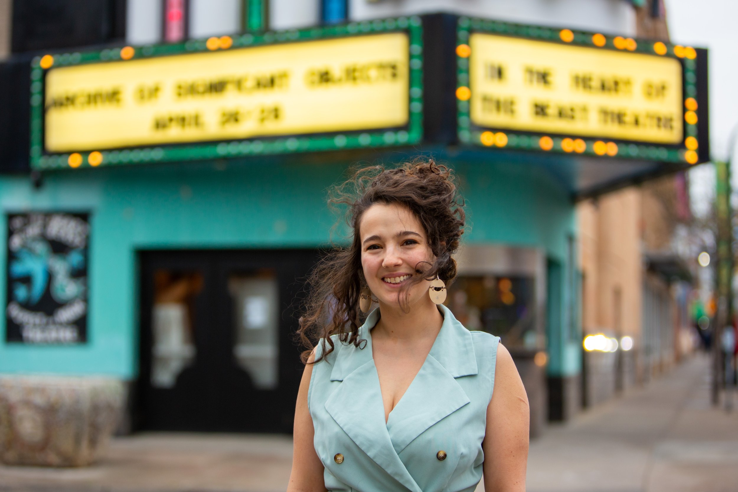 A smiling woman with curly brown hair wearing large circular earrings and a light blue sleeveless blazer standing on a city sidewalk in front of a theater marquee with yellow signs.