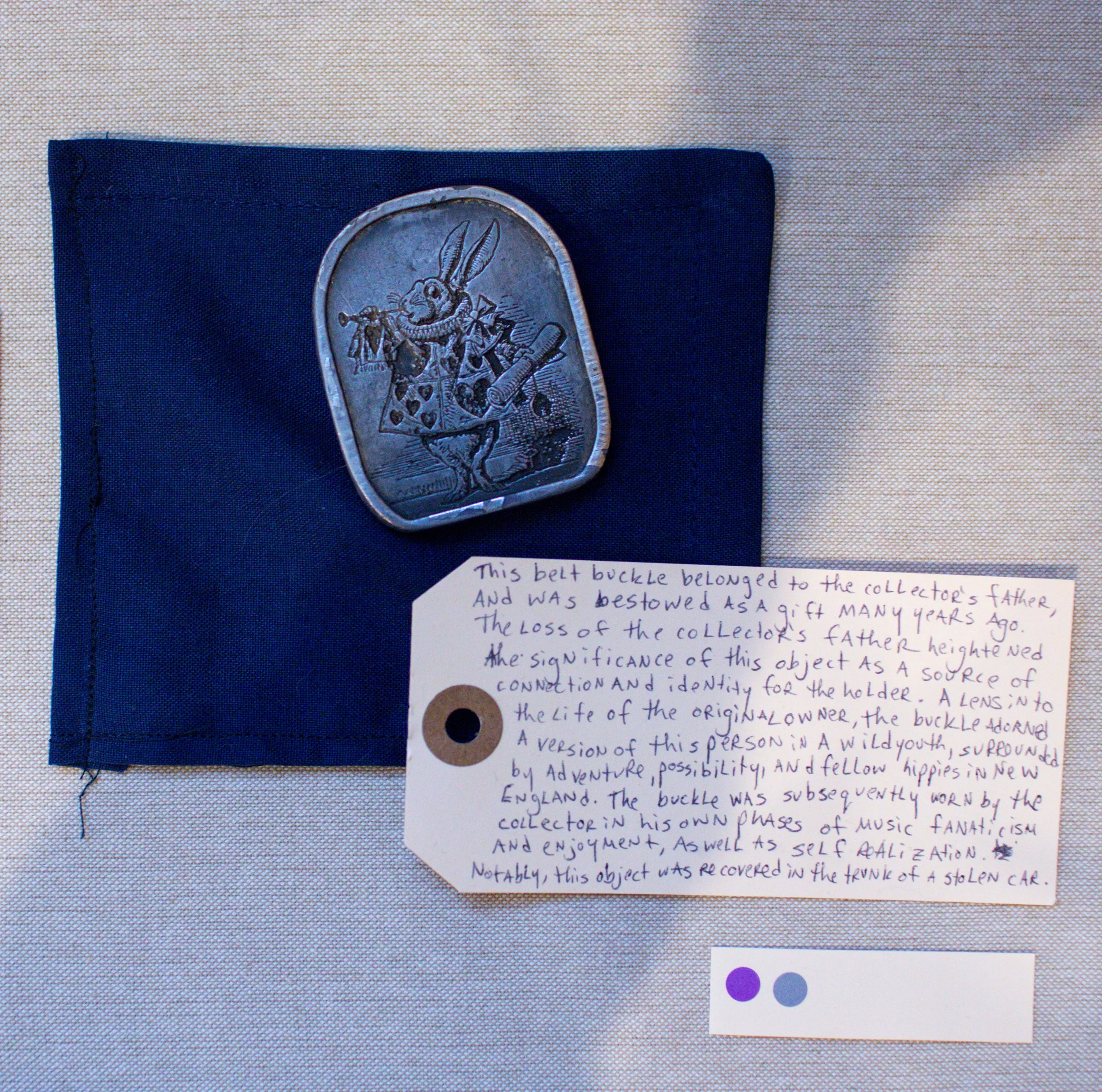 A silver belt buckle with a rabbit playing a flute, attached to a dark blue fabric background. A handwritten note explains that the buckle belonged to a collector's father, was a gift many years ago, and symbolizes connection and identity for the hol