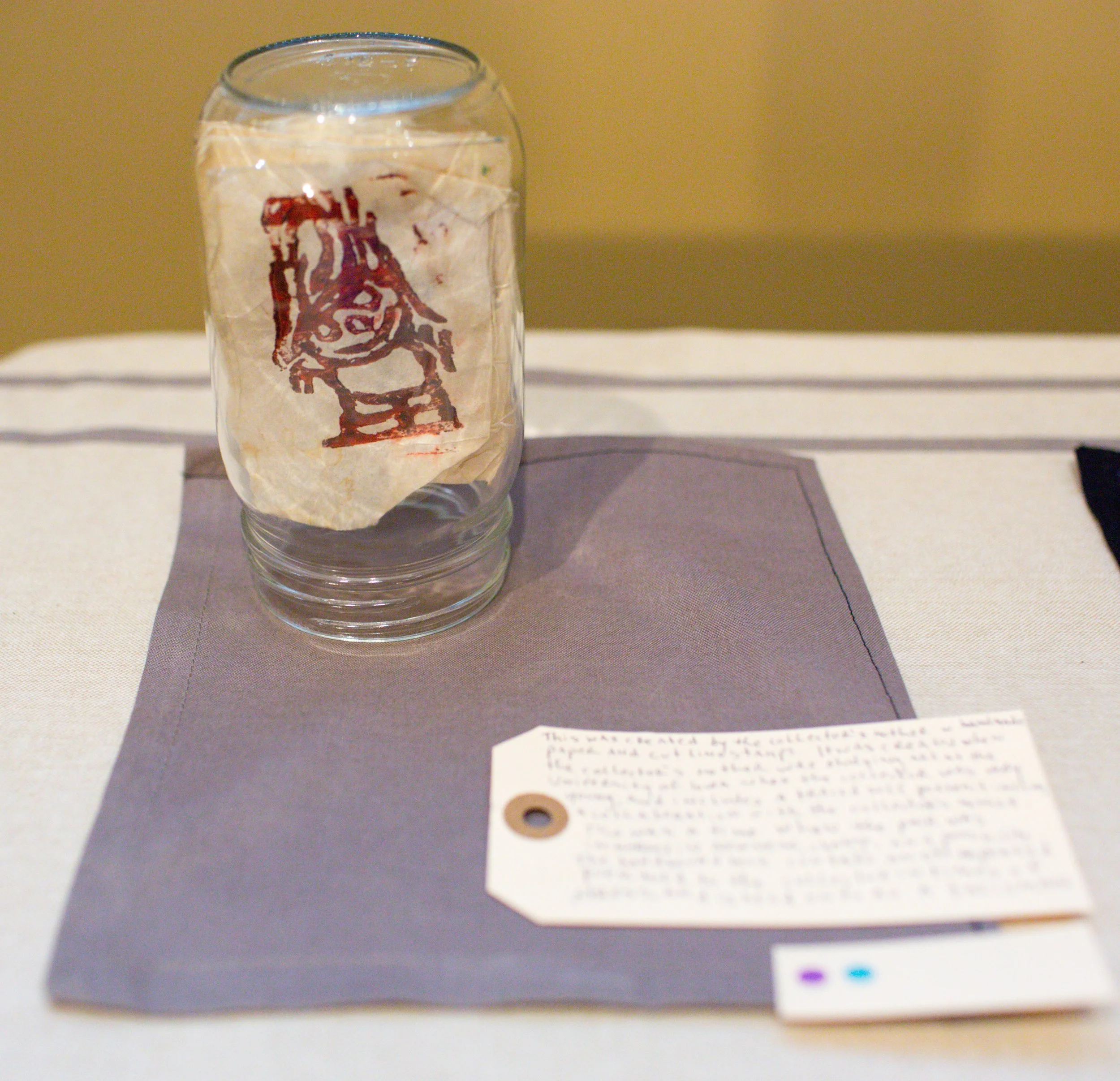 A clear glass with a drawing of a girl on a piece of tissue paper inside, placed on a gray cloth on a table.