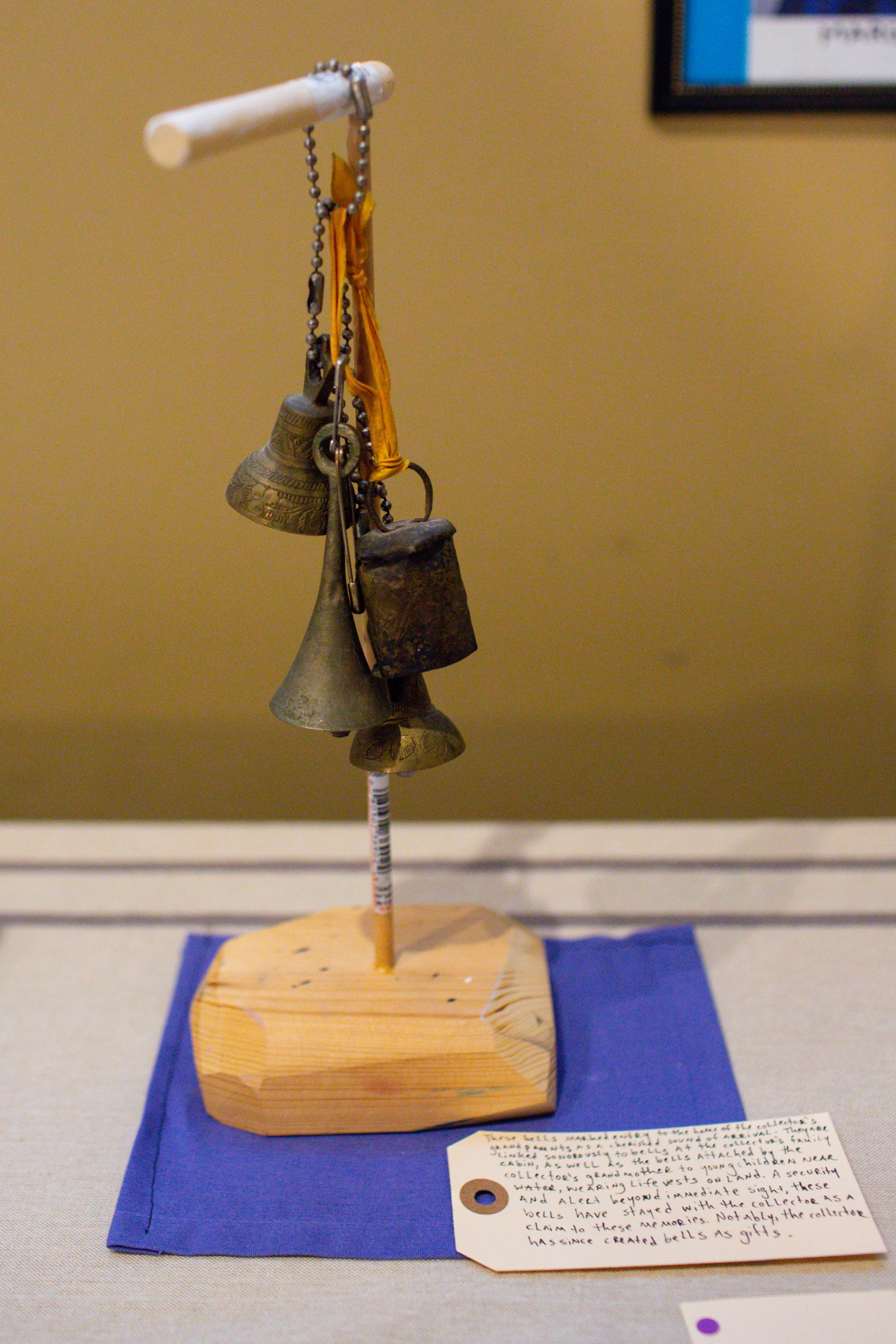 A small display of vintage metal bells hanging from a string on a white wooden stand, placed on a blue cloth with a handwritten description card.
