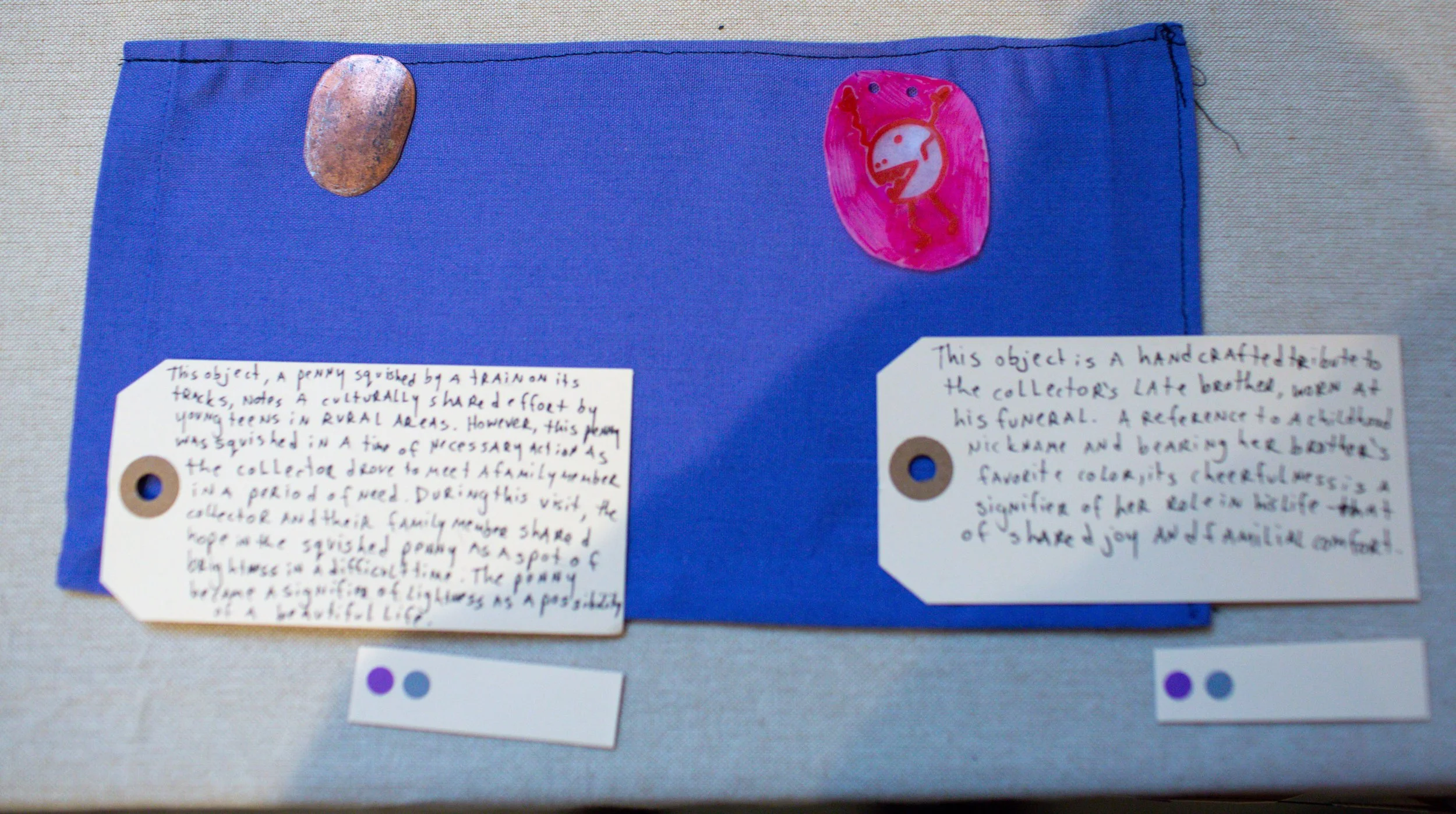 Handmade blue fabric with two painted rocks, one with a copper color and the other with a pink and white ladybug drawing. Two handwritten notes on white paper tags describe the objects.