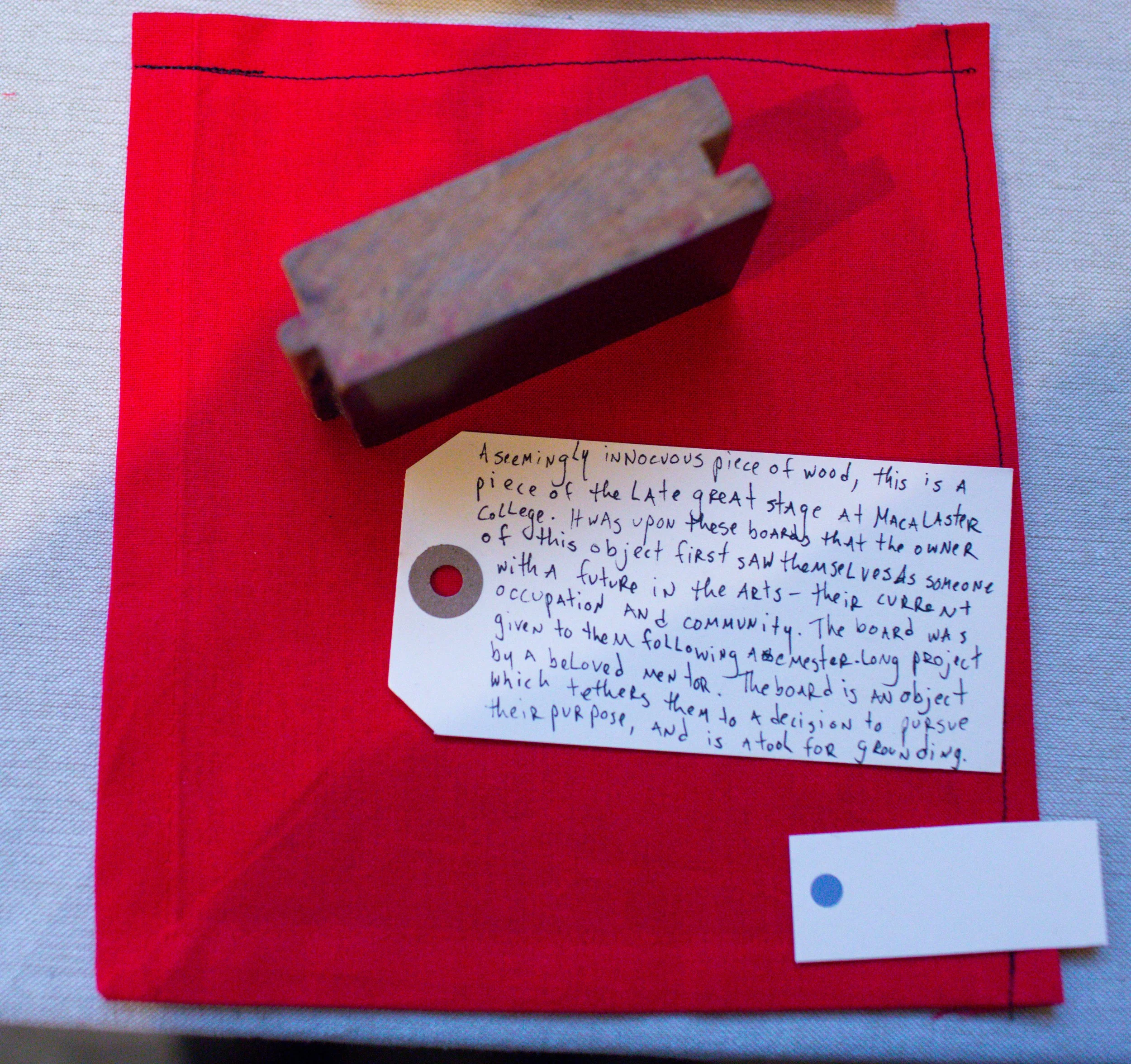 A wooden stamp and a handwritten note on red fabric with a white background. The note explains that the stamp is a piece of a late great stage at Macalaster College, given to the owner by a board member to symbolize a future in the arts and community