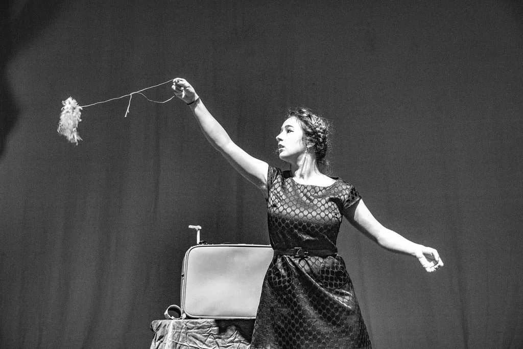 A woman in a patterned dress holding a feather duster on a stage with a suitcase on a stand nearby.