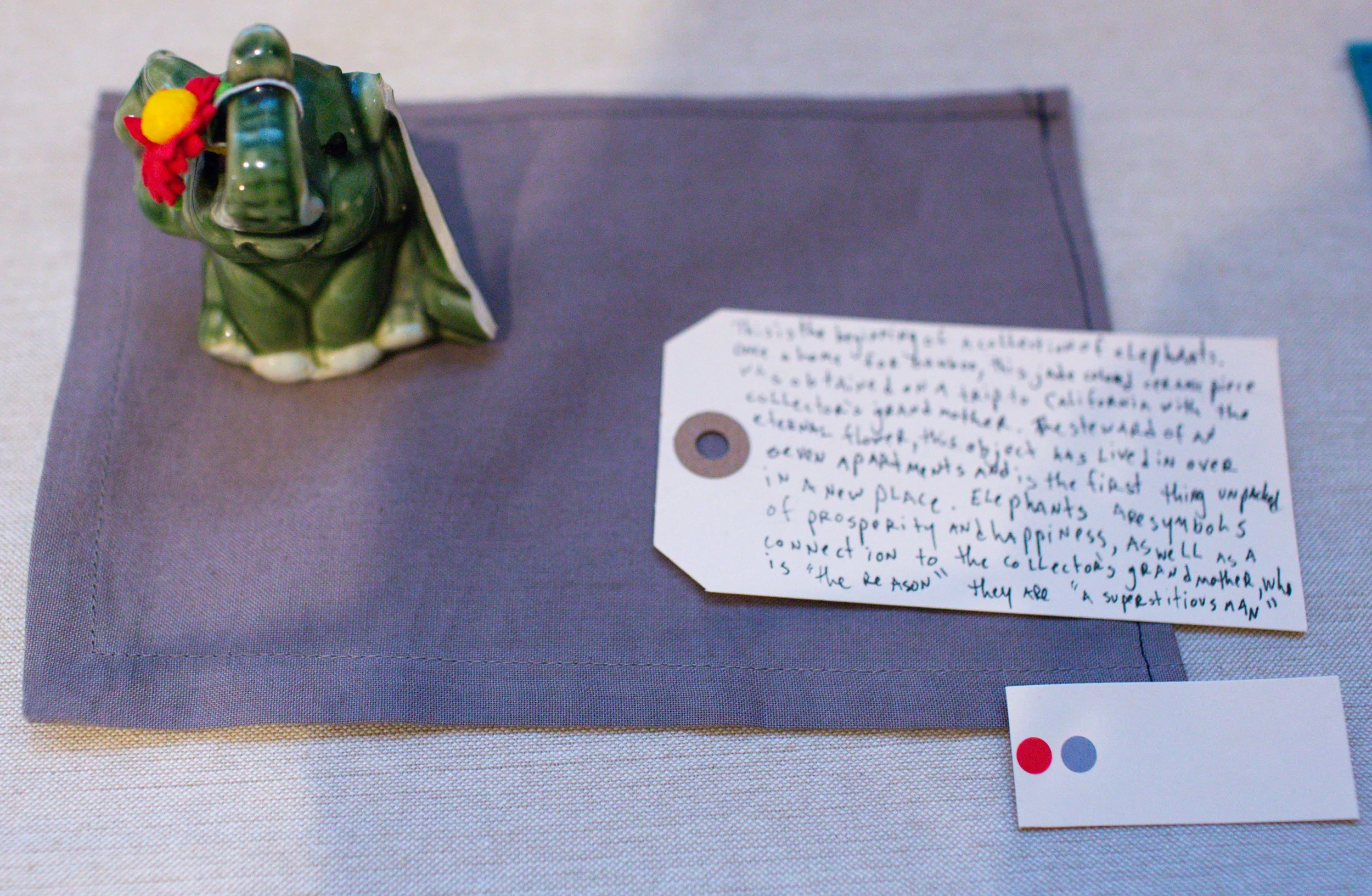 A ceramic frog figurine with a red and yellow flower on its head, sitting on a purple cloth placemat. Next to it is a handwritten note on a white tag describing the frog and its symbolism, and a small white card with two colored dots, red and gray.