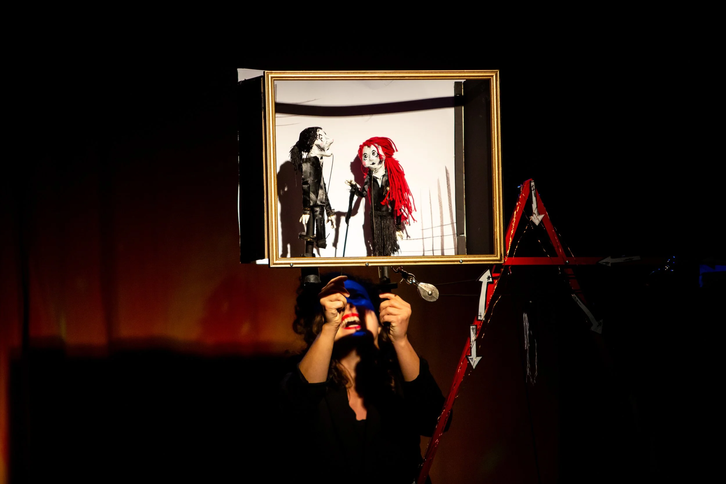 A puppeteer performing a puppet show with two marionette characters, a man and a woman, inside a gold frame, with the puppeteer unseen behind the setup, holding the controls.