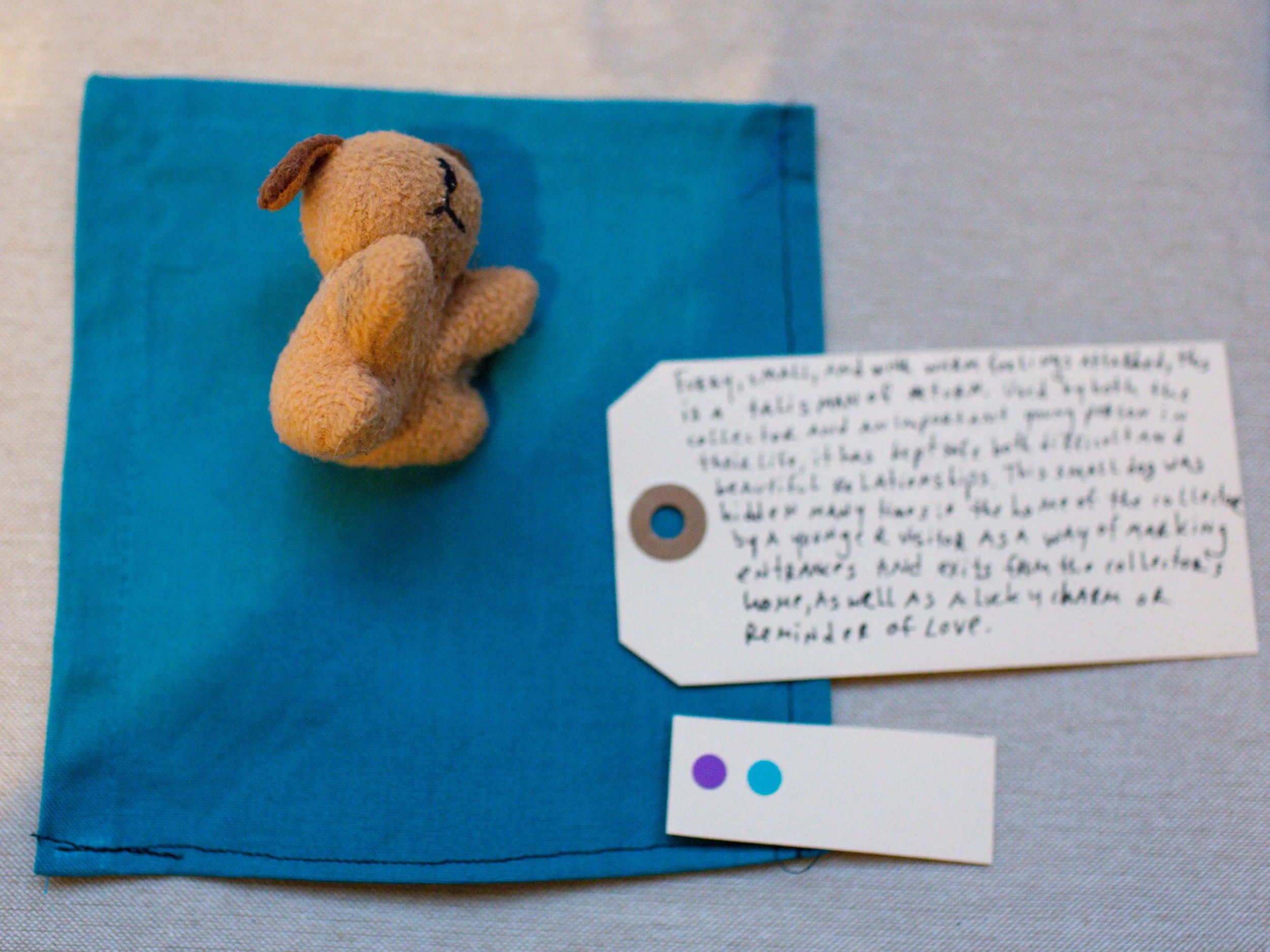 A small plush bear lying on a blue fabric square with a white handwritten note and a small white label with purple and blue dots.