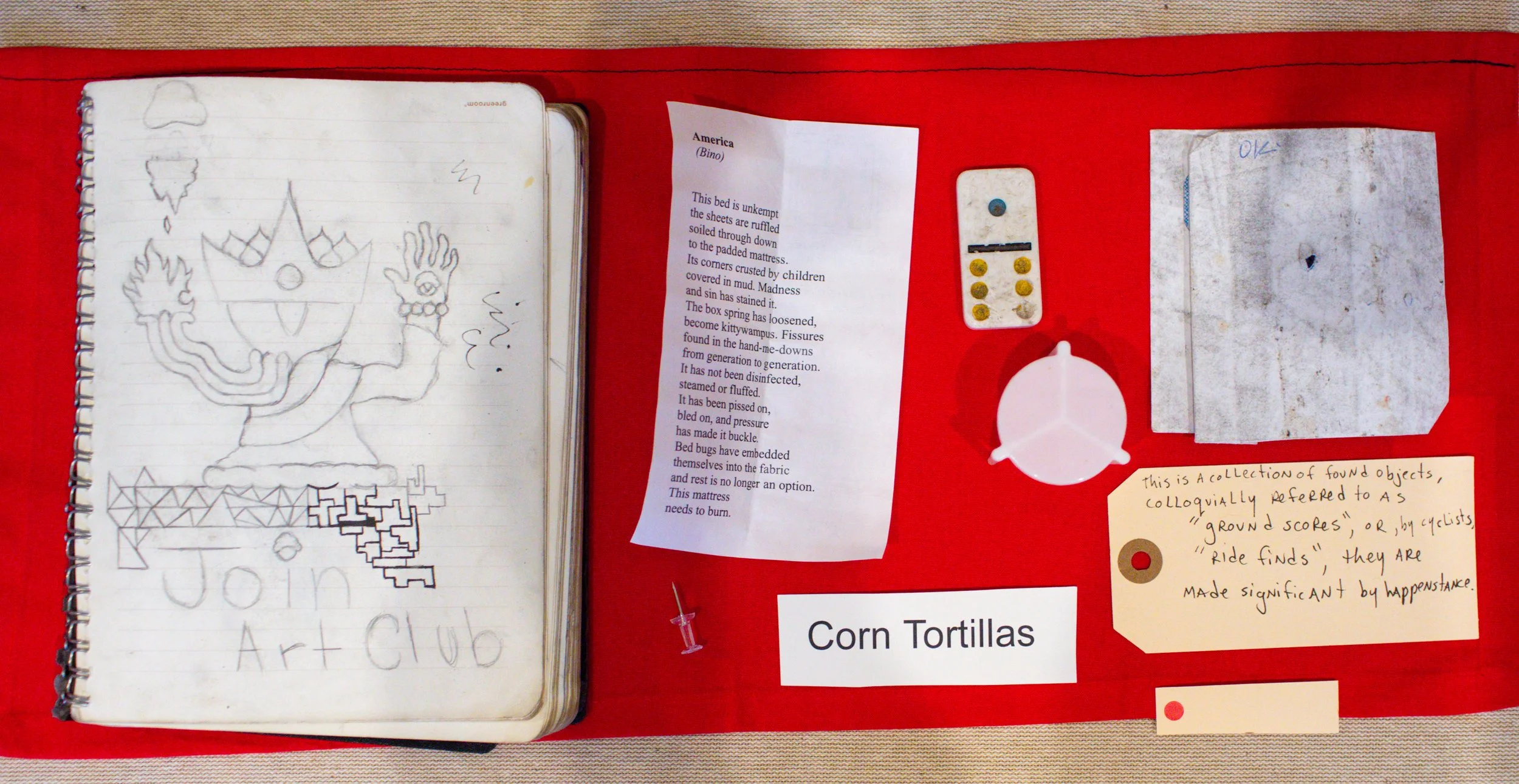Display of a red board with various items related to a project, including a notebook with a handwritten drawing and the words 'Join Art Club,' a printed poem about a bed, a small container with yellow and blue objects, a crumpled piece of paper, a sm