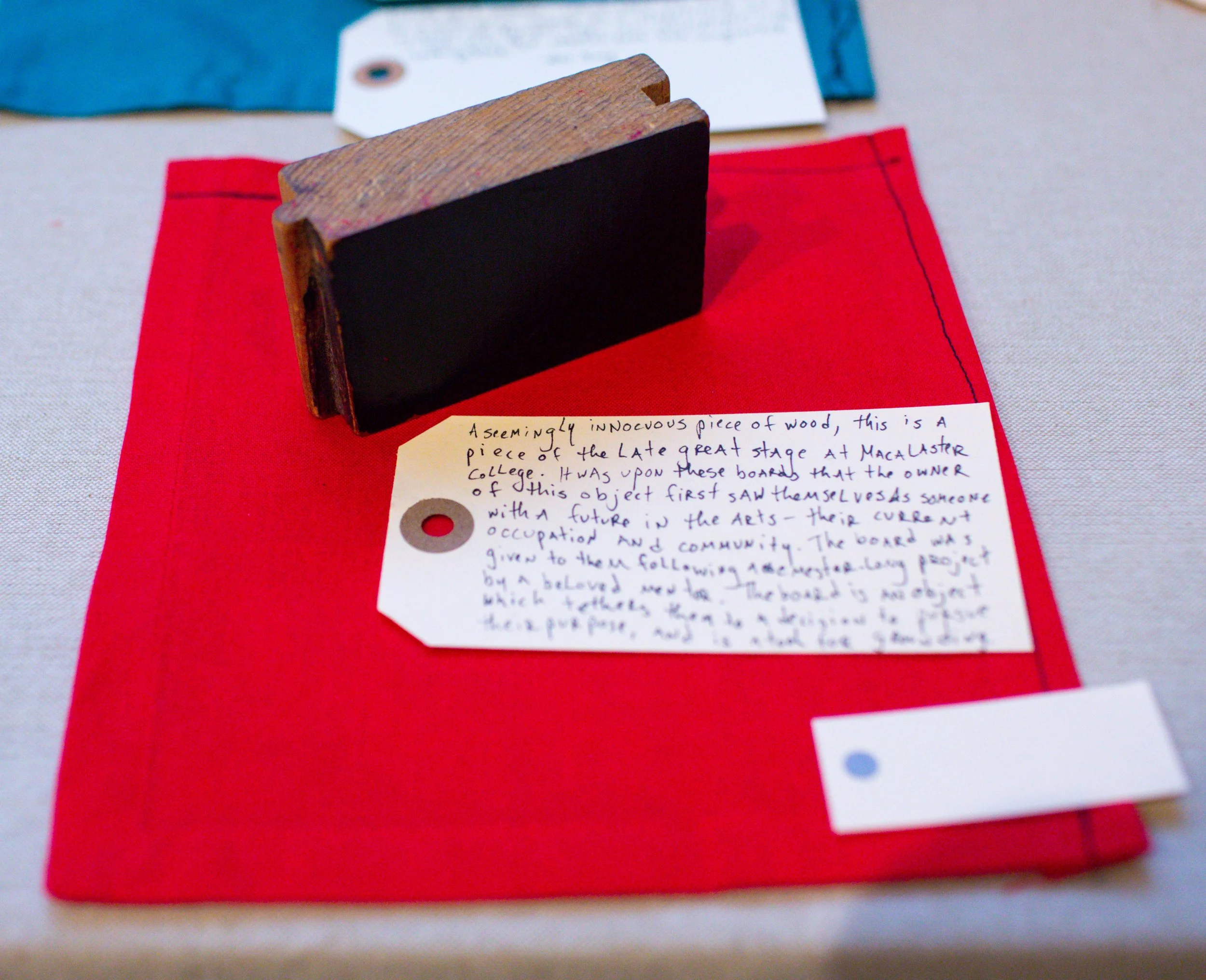 A small wooden blackboard with a black surface, placed on a red cloth with a white paper tag attached, surrounded by other cloths of different colors.
