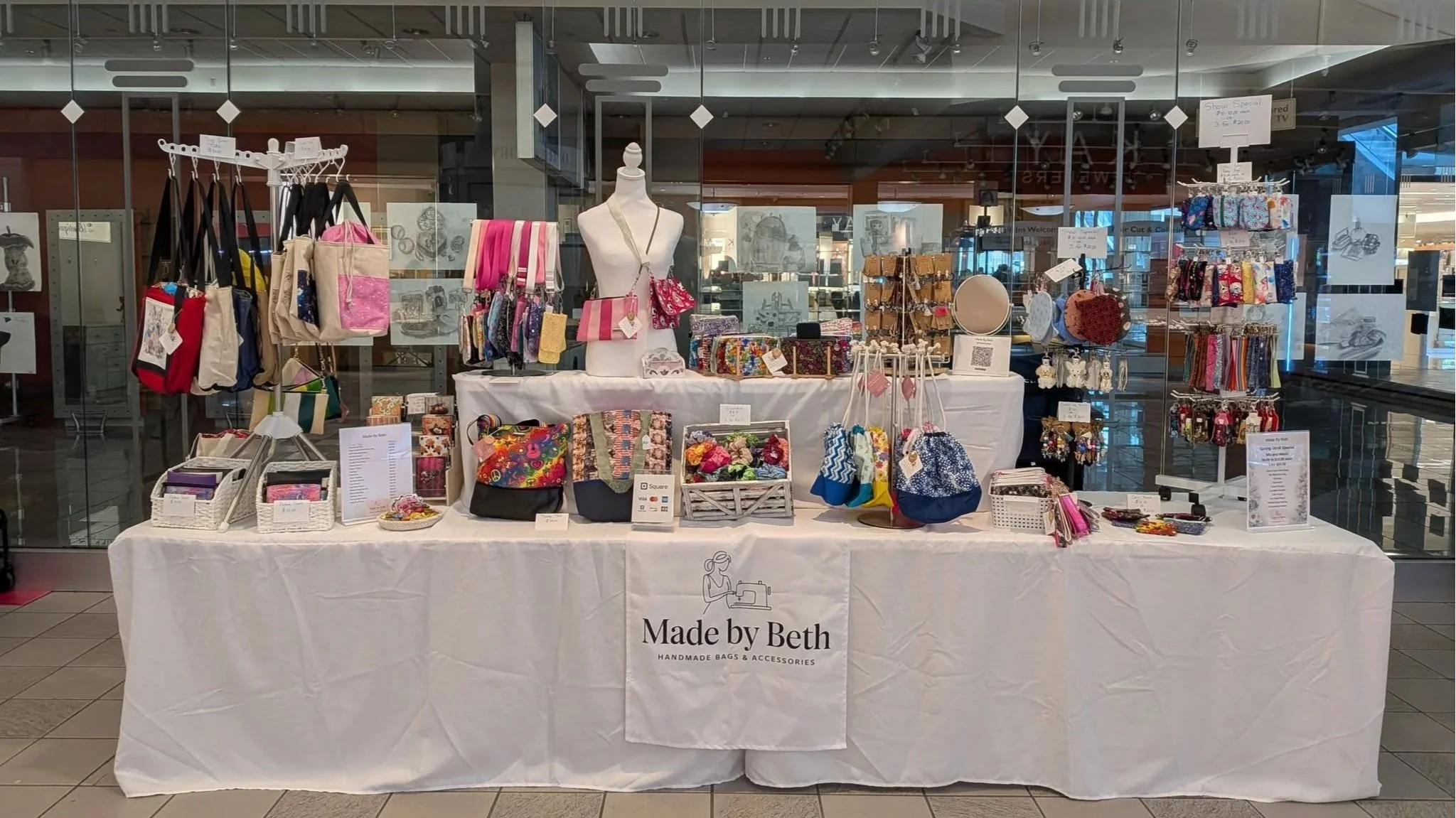 A display table with handmade bags and accessories at a craft fair, organized with various bags, jewelry, and small decorative items, labeled 'Made by Beth'.