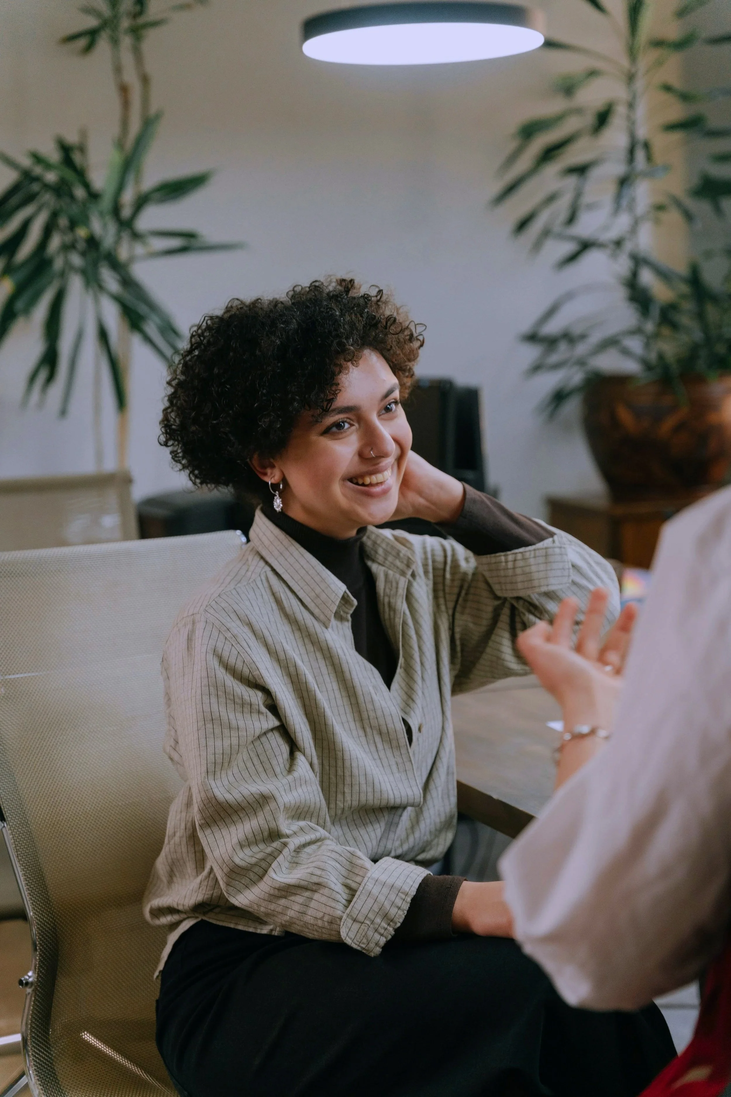 A young woman with curly hair, wearing a beige striped shirt and dark pants, smiling and engaged in conversation with someone whose hand is gesturing. She is seated indoors with plants and a ceiling light in the background.