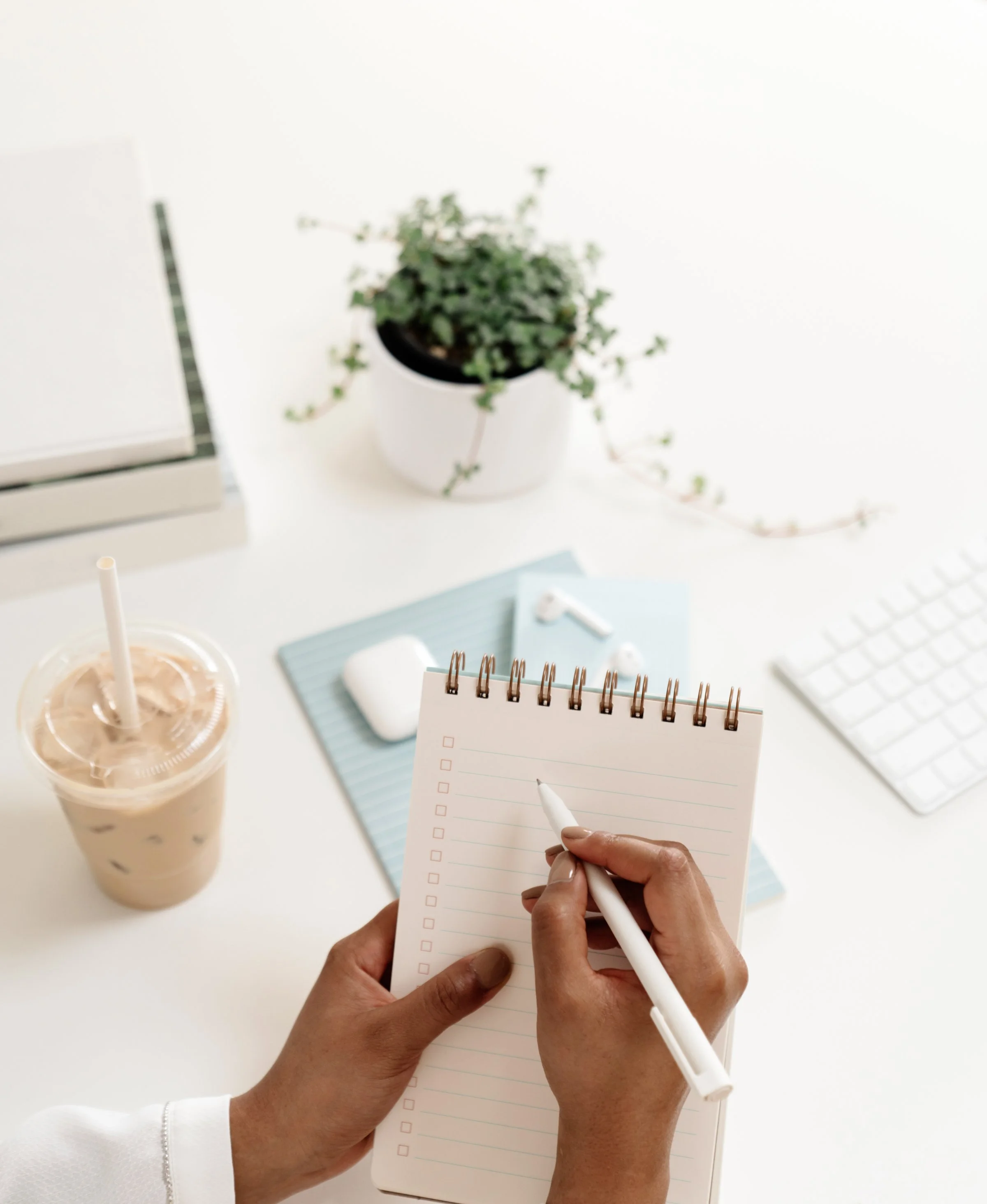 A person holding a notepad and writing with a white pen on a white desk with various office items, including a plant, coffee drink, wireless earbuds, a keyboard, and notebooks.