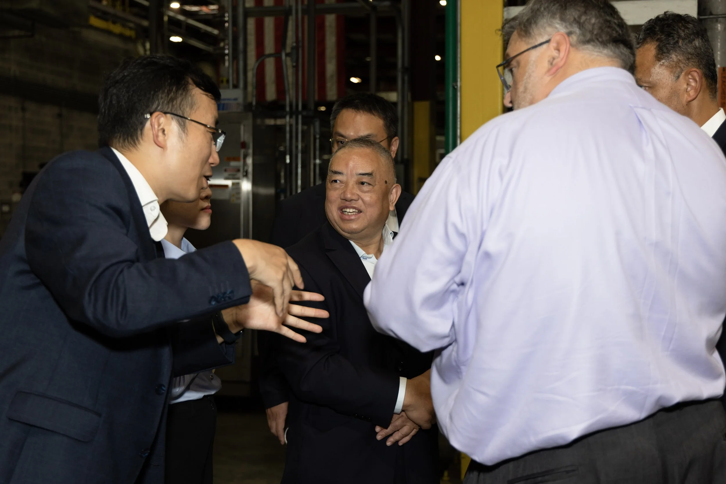 Group of men engaged in conversation, with one man in a suit speaking and others listening, in an industrial or warehouse setting.