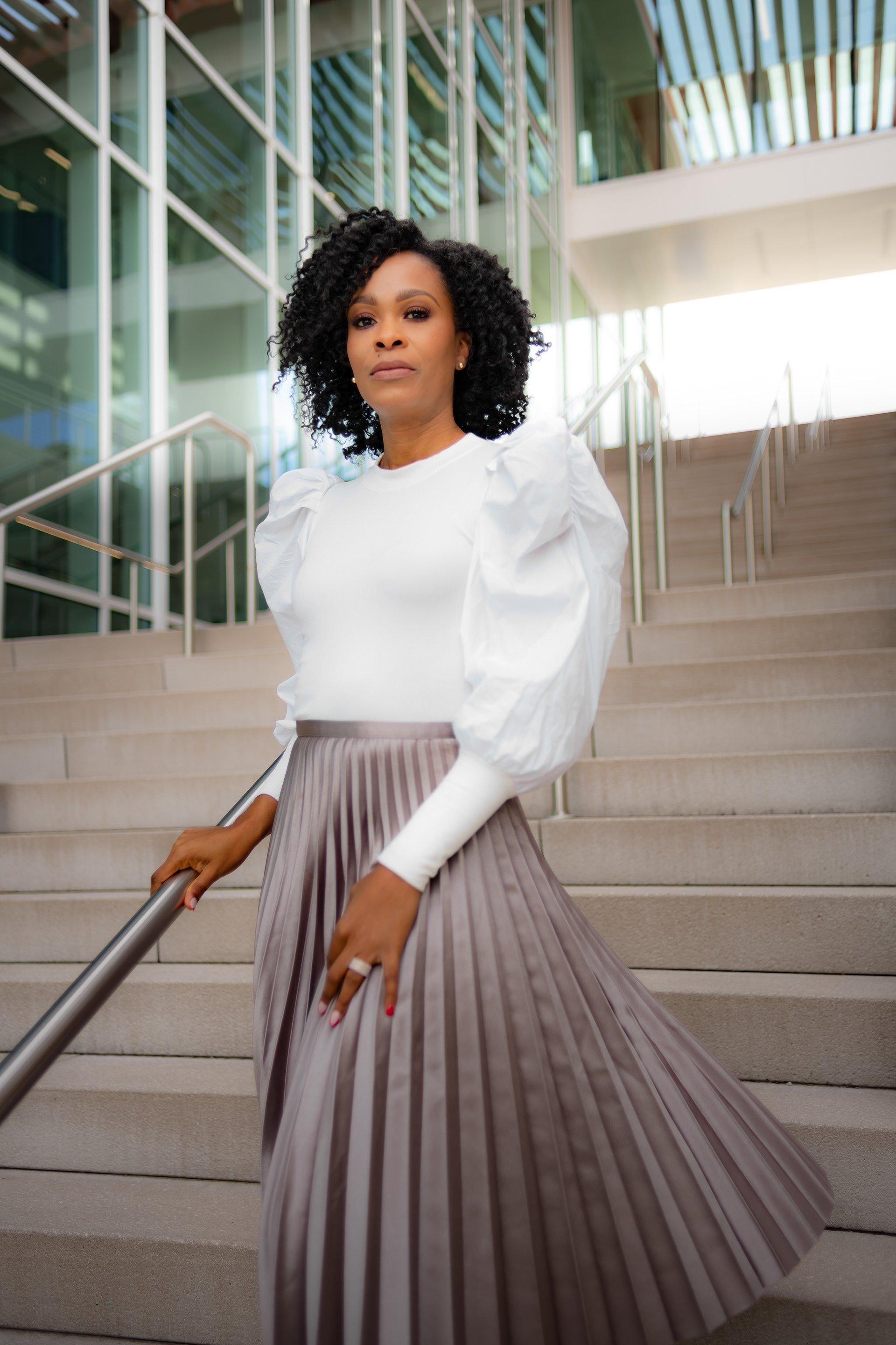 A woman with curly black hair stands on a staircase outside a modern glass building, wearing a white blouse with puffed sleeves and a pleated gray skirt.